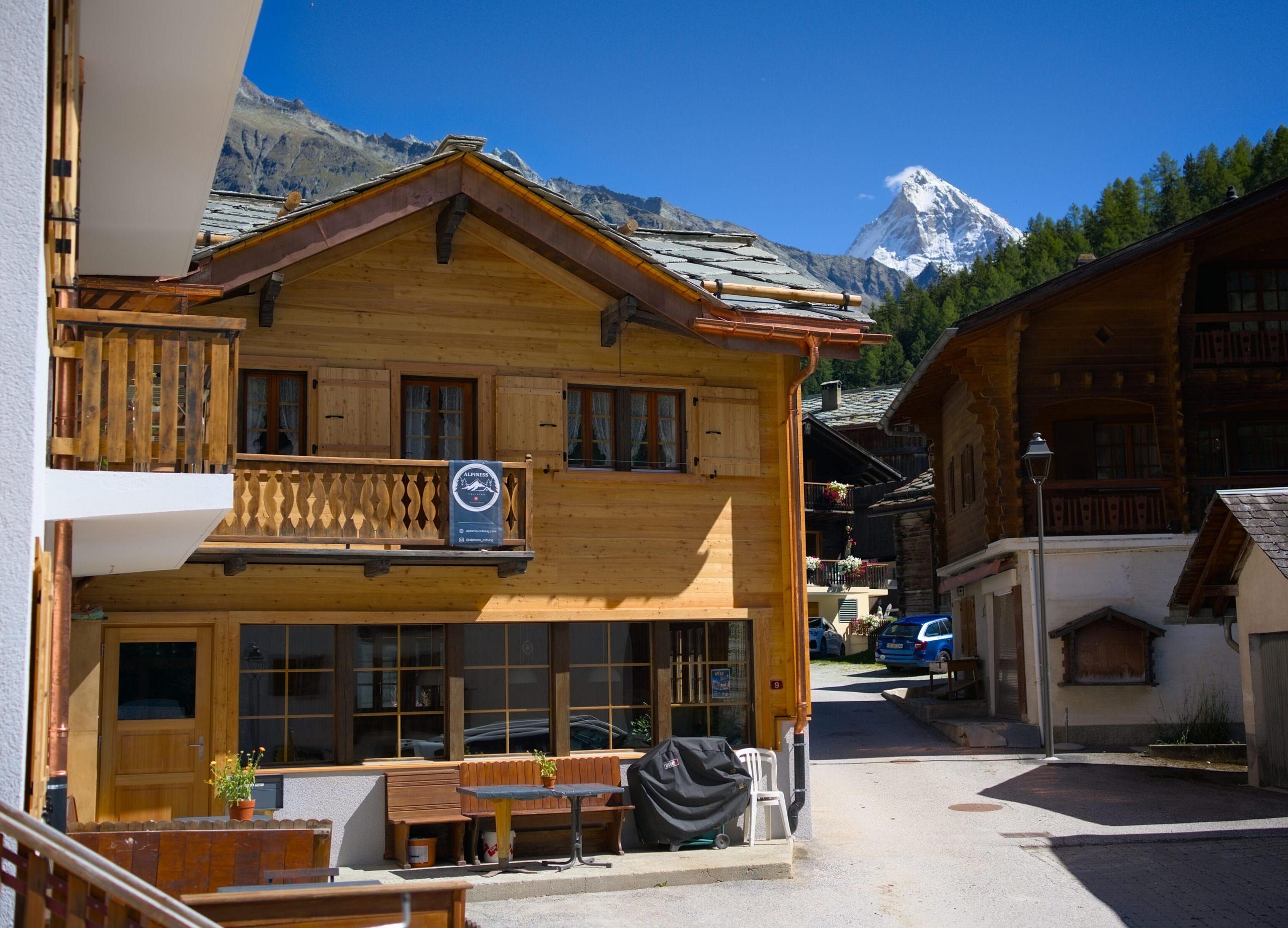 Side view of the Alpiness coliving building with the Dent Blanche peak visible in the background, showing the renovated wooden facade and the outdoor entrance area.