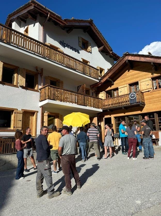 A group of people gathered outside the Alpiness coliving building for an apéro, with the renovated white facade and Alpiness sign visible in the background.