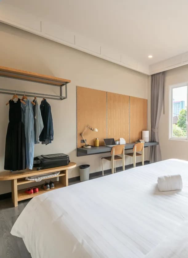 Spacious room with a large white bed, wooden desk with two chairs, open wardrobe with hanging clothes, and a window with natural light