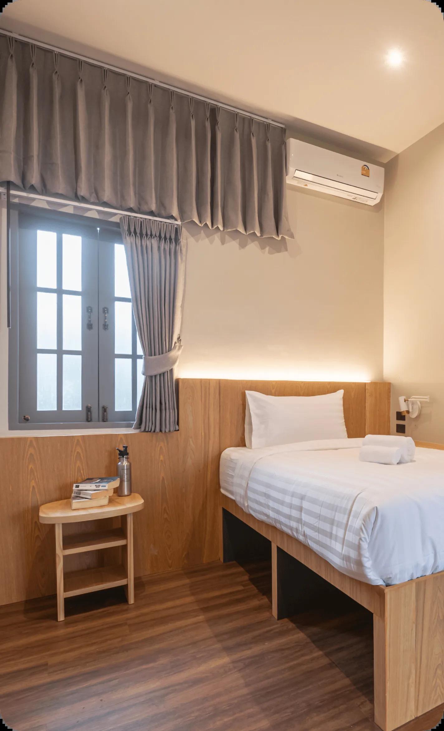 Small single room with a single bed, wooden frame, side table, window with curtains, and air conditioning unit