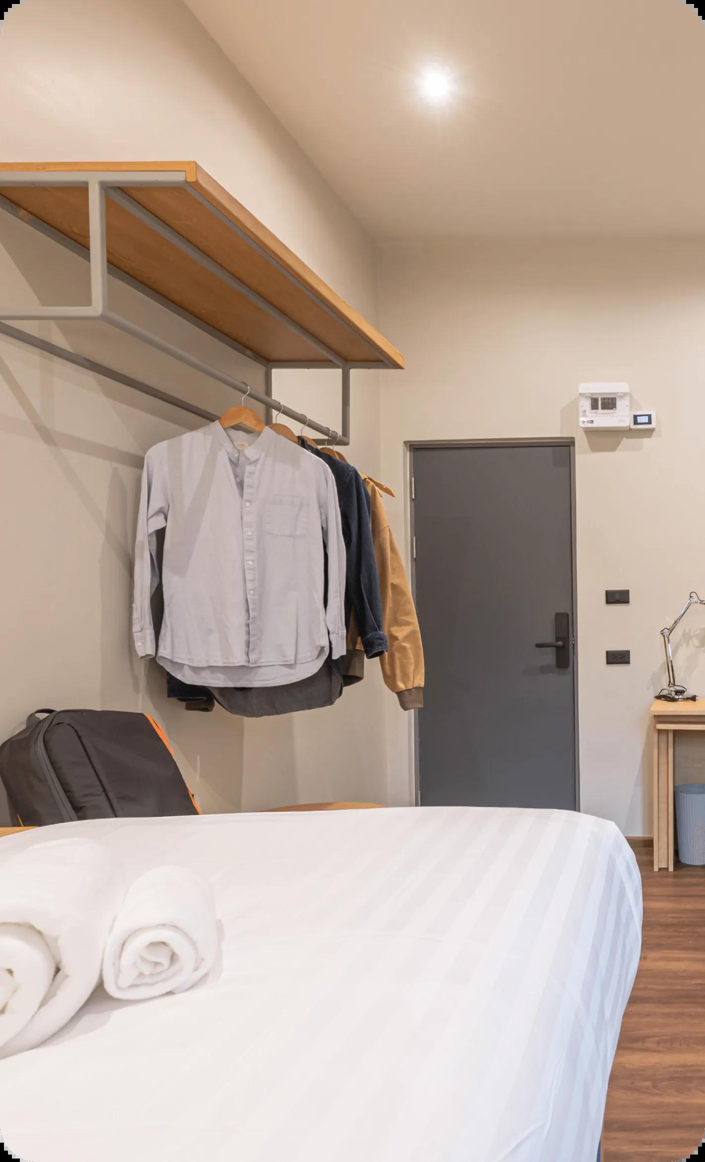 Room corner showing open wardrobe rack with hanging clothes, towels on bed, and grey door