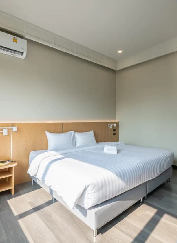 Bright room with a large king-size bed with white linens, wooden headboard panel, wall-mounted reading light, and air conditioning unit