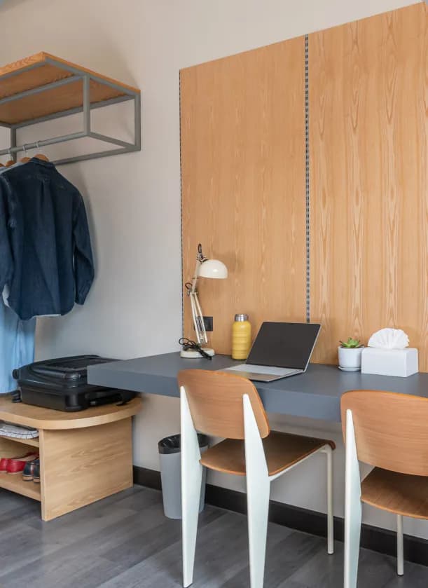 Room desk area with a long grey desk, two wooden chairs, open wardrobe with hanging clothes, and a suitcase on a bench
