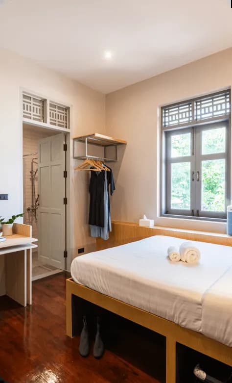 Cozy room with a single bed with white linens, open wardrobe with hanging clothes, window with garden view, and en-suite bathroom door