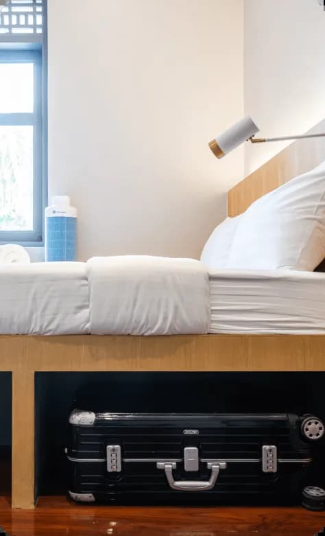 Bed with white linens showing under-bed suitcase storage space, with a bedside lamp and window