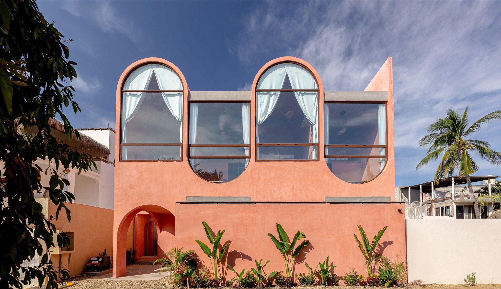 Front facade of Amplitude Coliving building in La Punta, Puerto Escondido — a terracotta-pink building with distinctive arched windows and tropical plants at the entrance