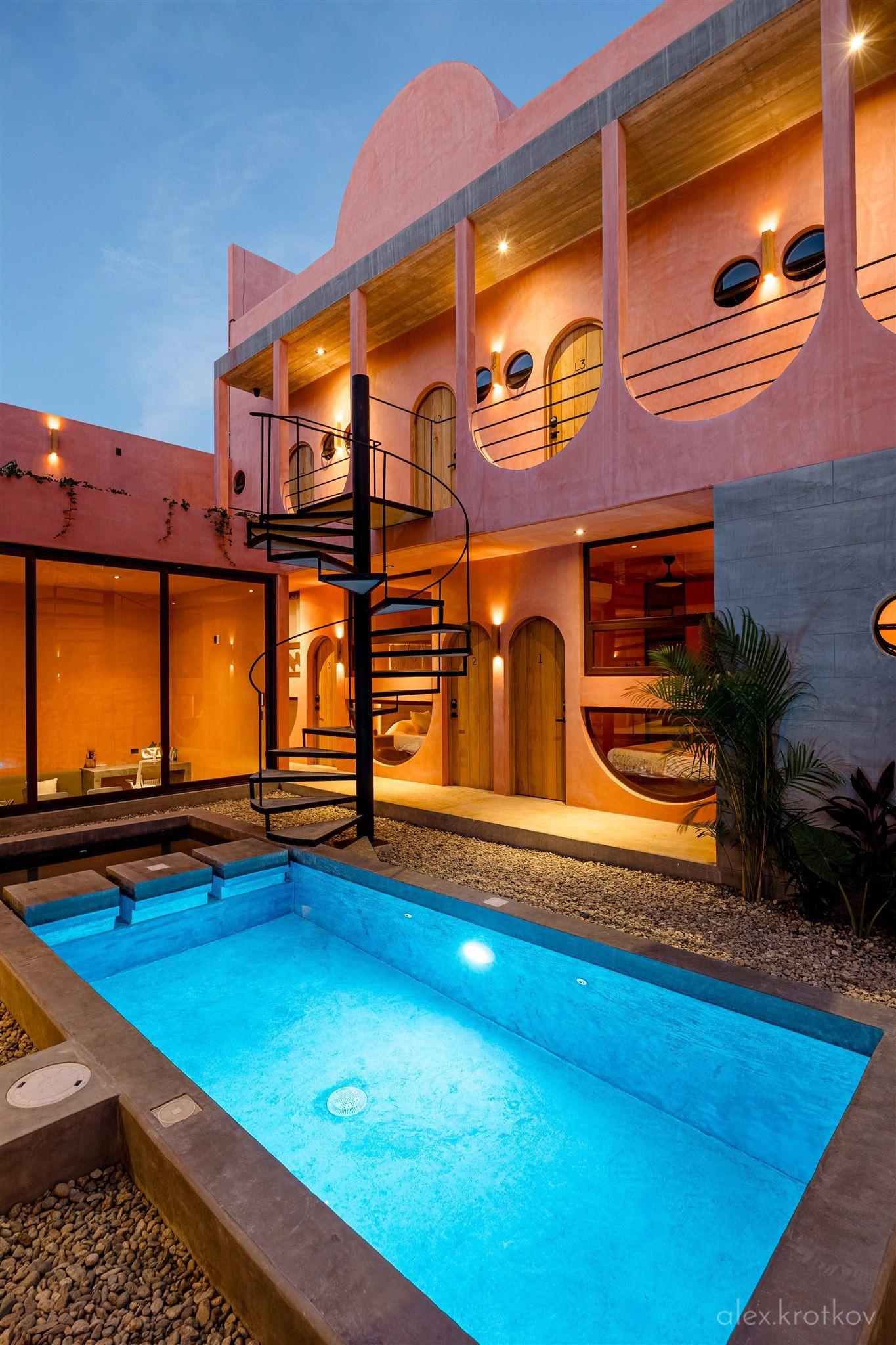 Outdoor pool and courtyard of Amplitude Coliving at dusk, showing the illuminated building facade, spiral staircase, and glowing blue pool