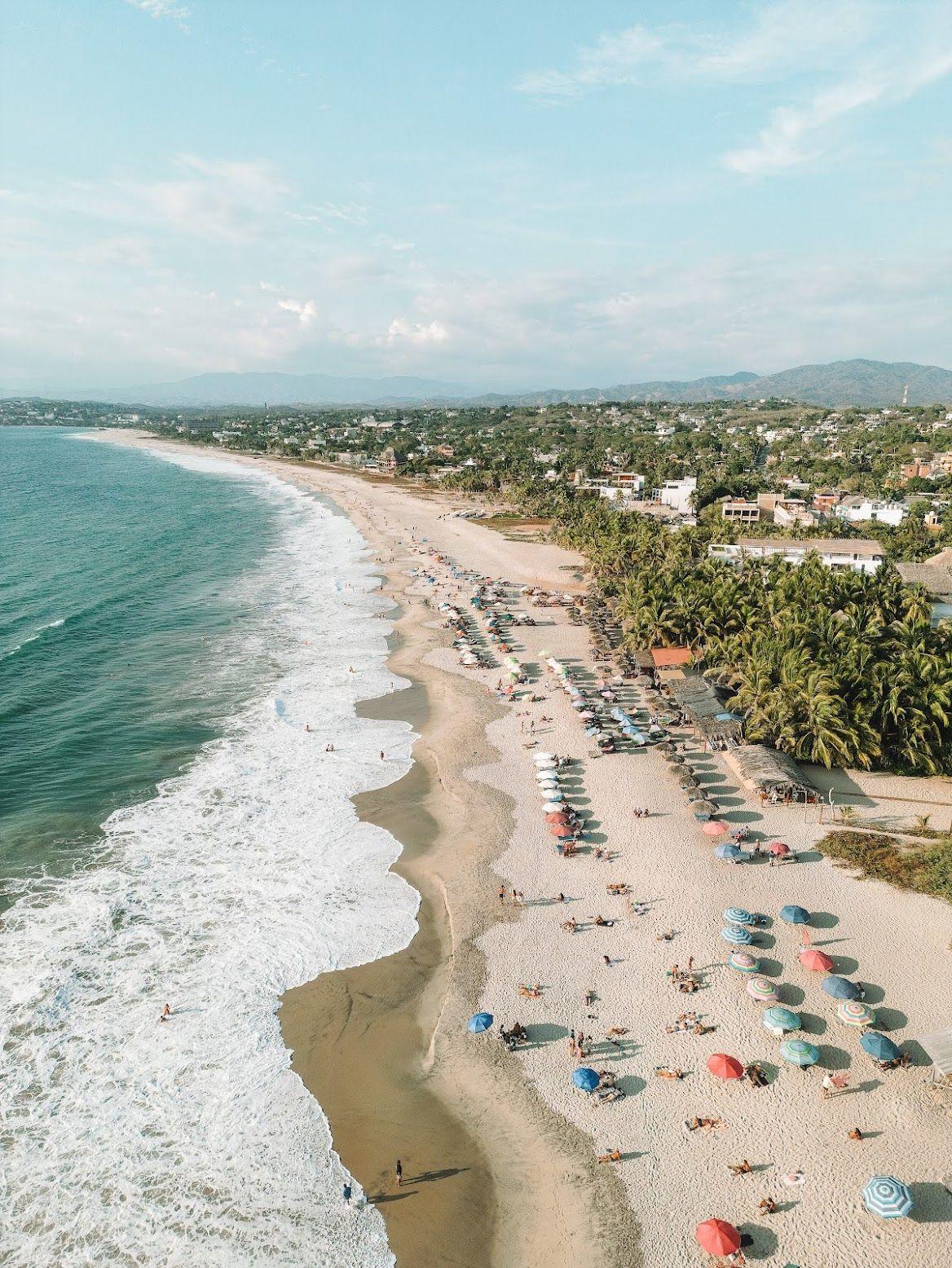 Aerial view of a beach in Puerto Escondido, showing the coastline, surf, beach umbrellas, and the town in the background