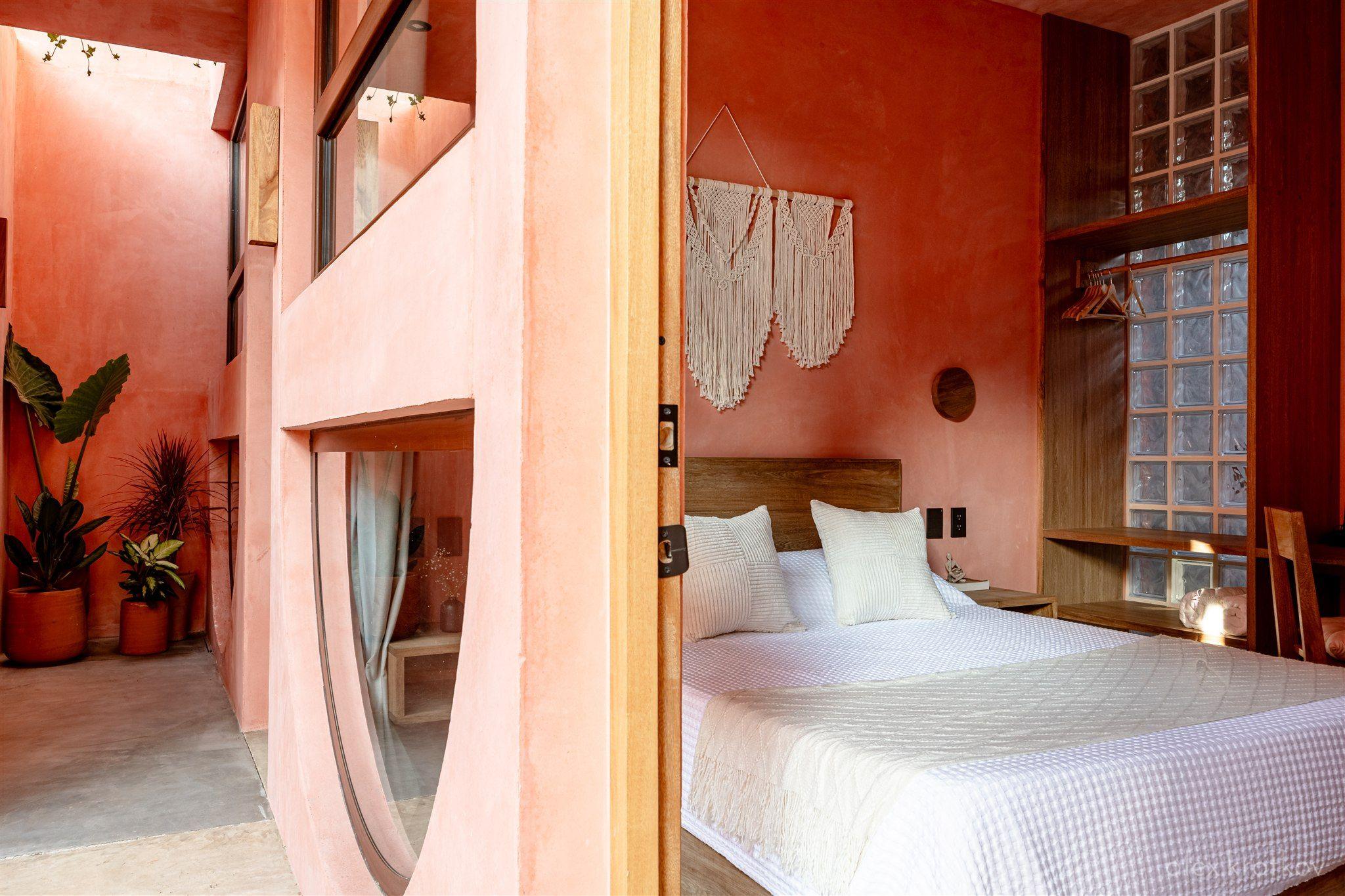 Queen room at Amplitude Coliving, showing a king bed with macramé wall hanging and view into the courtyard through an arched doorway