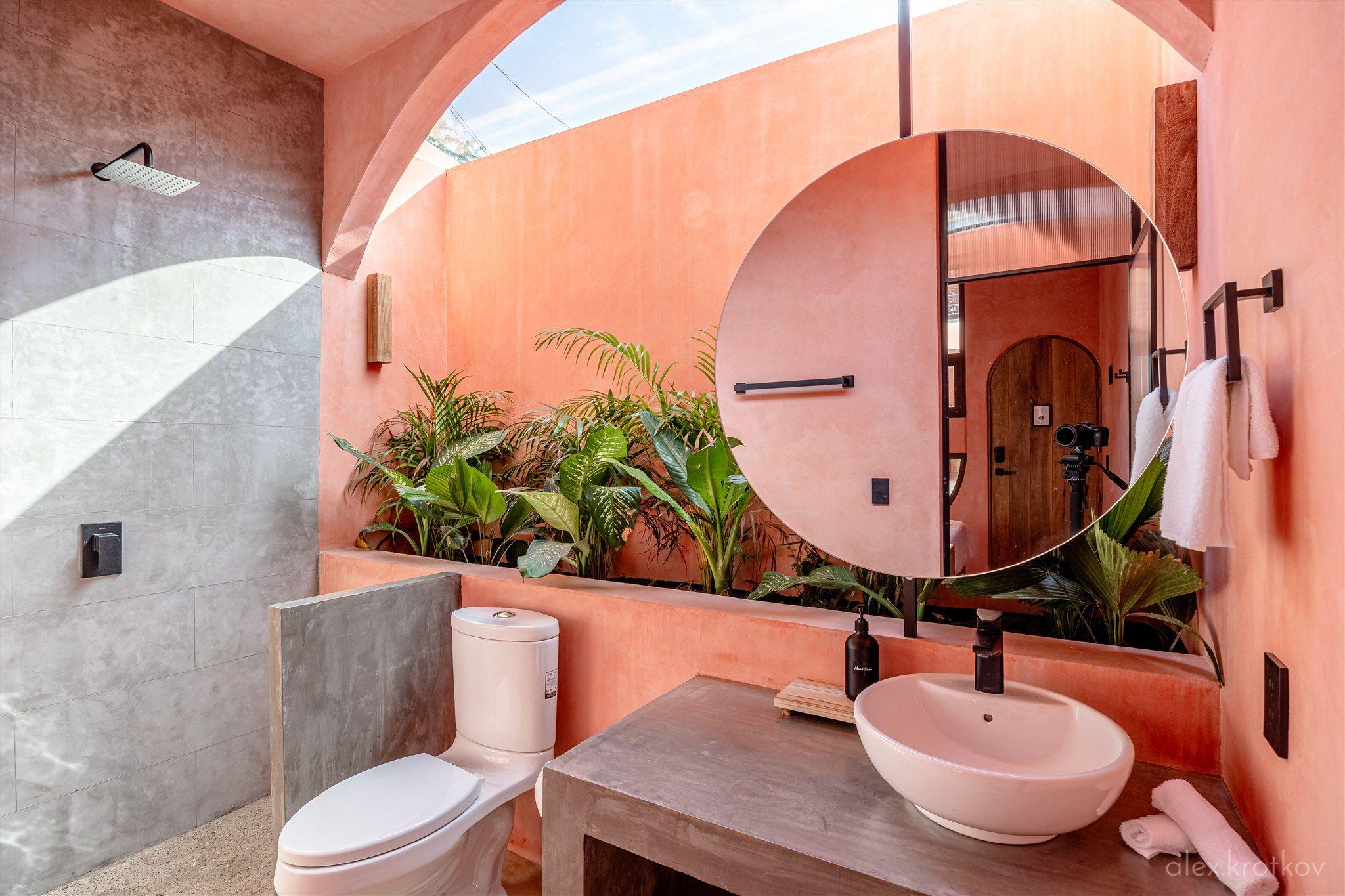 Ensuite bathroom of the Garden Bath Queen Room at Amplitude Coliving, featuring a glass ceiling, round mirror, vessel sink, and lush plants visible through an arched opening