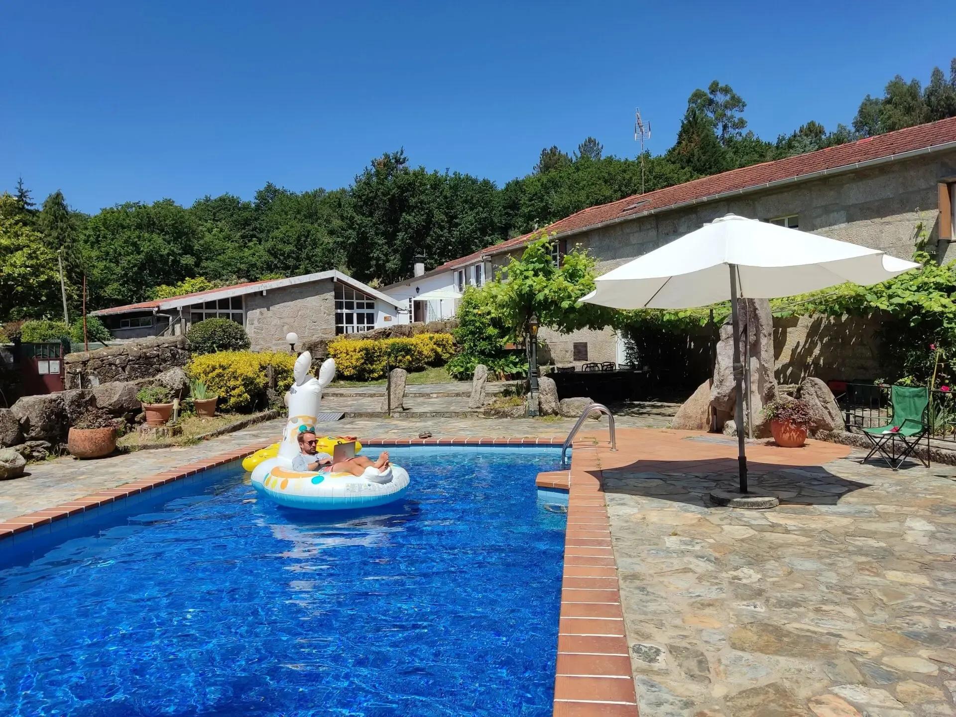 Outdoor swimming pool with a person floating on an inflatable, surrounded by the coliving buildings and garden