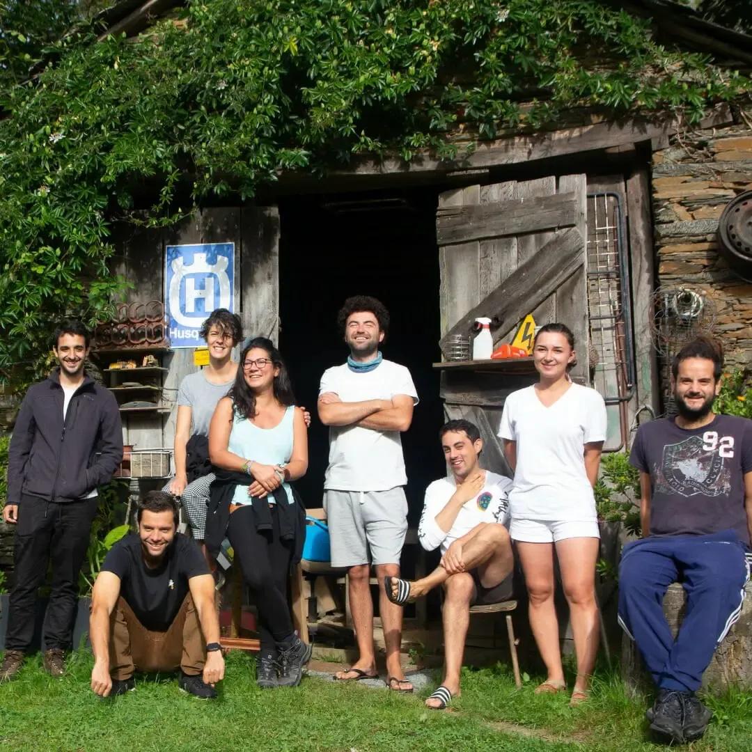 Group of eight people posing in front of a rustic stone building with a Husqvarna sign, in a rural garden setting