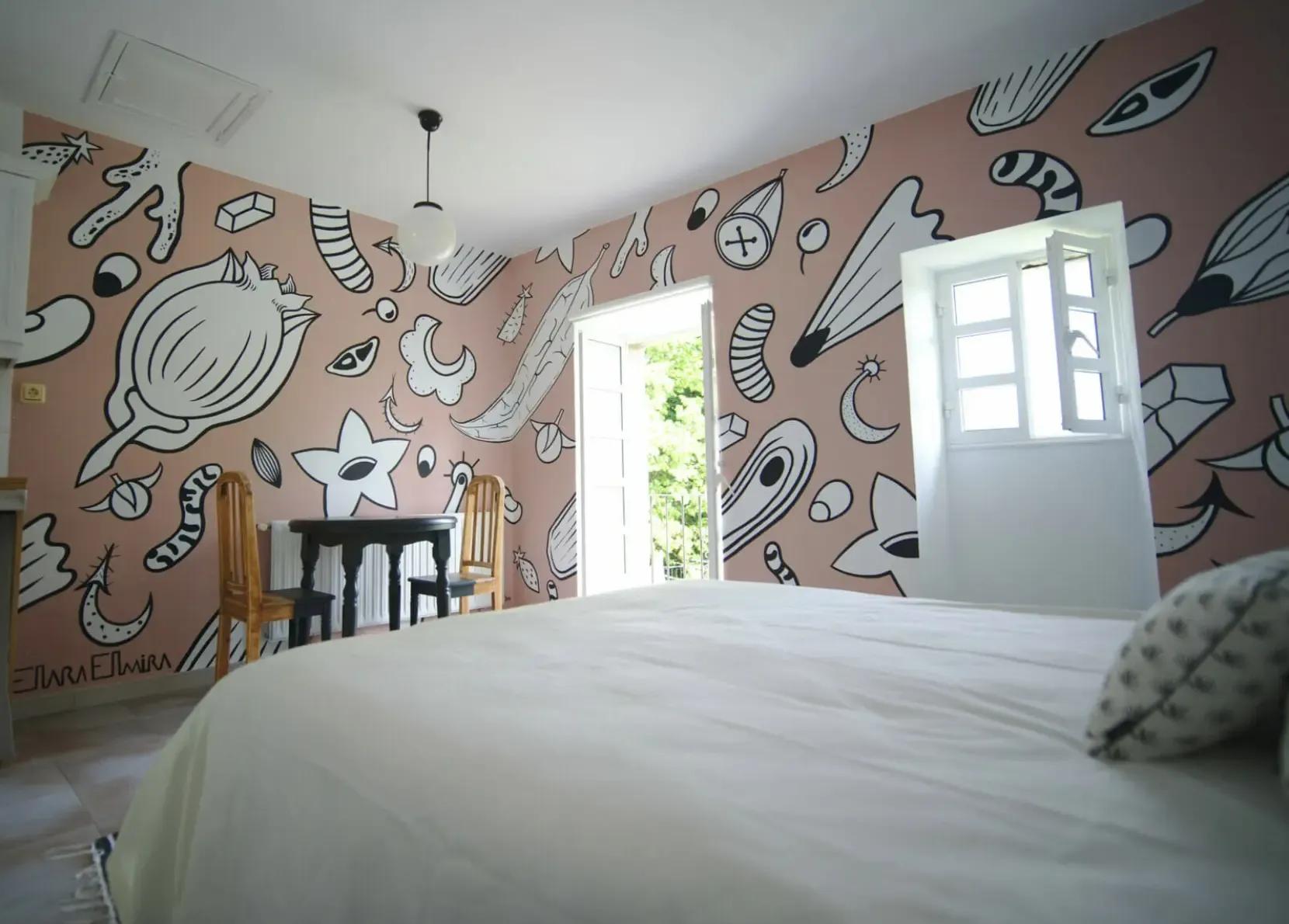 Private room with a large artistic mural on pink walls, white bed, desk and chairs, and a window with garden view