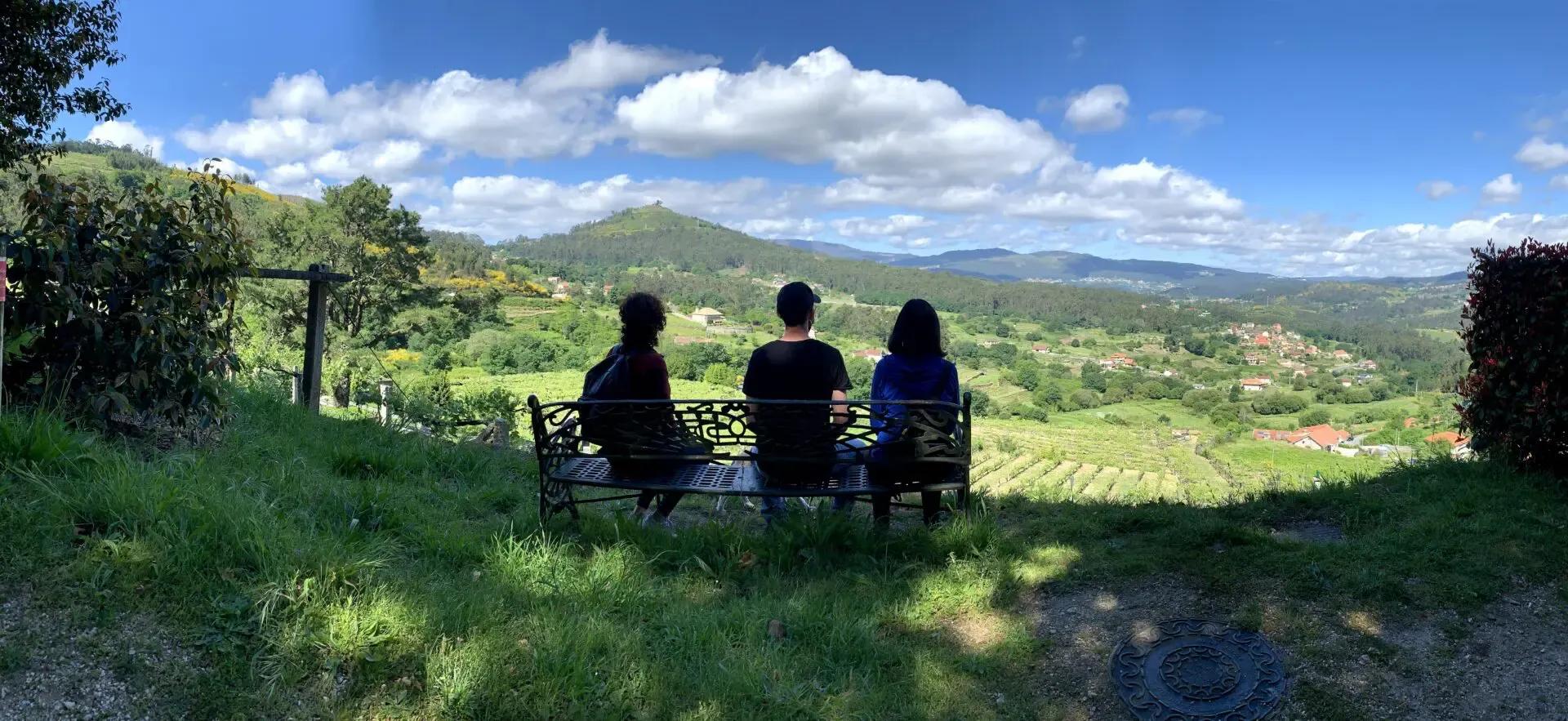 Three people sitting on a bench with their backs to the camera, looking out over the green Galician countryside and vineyards