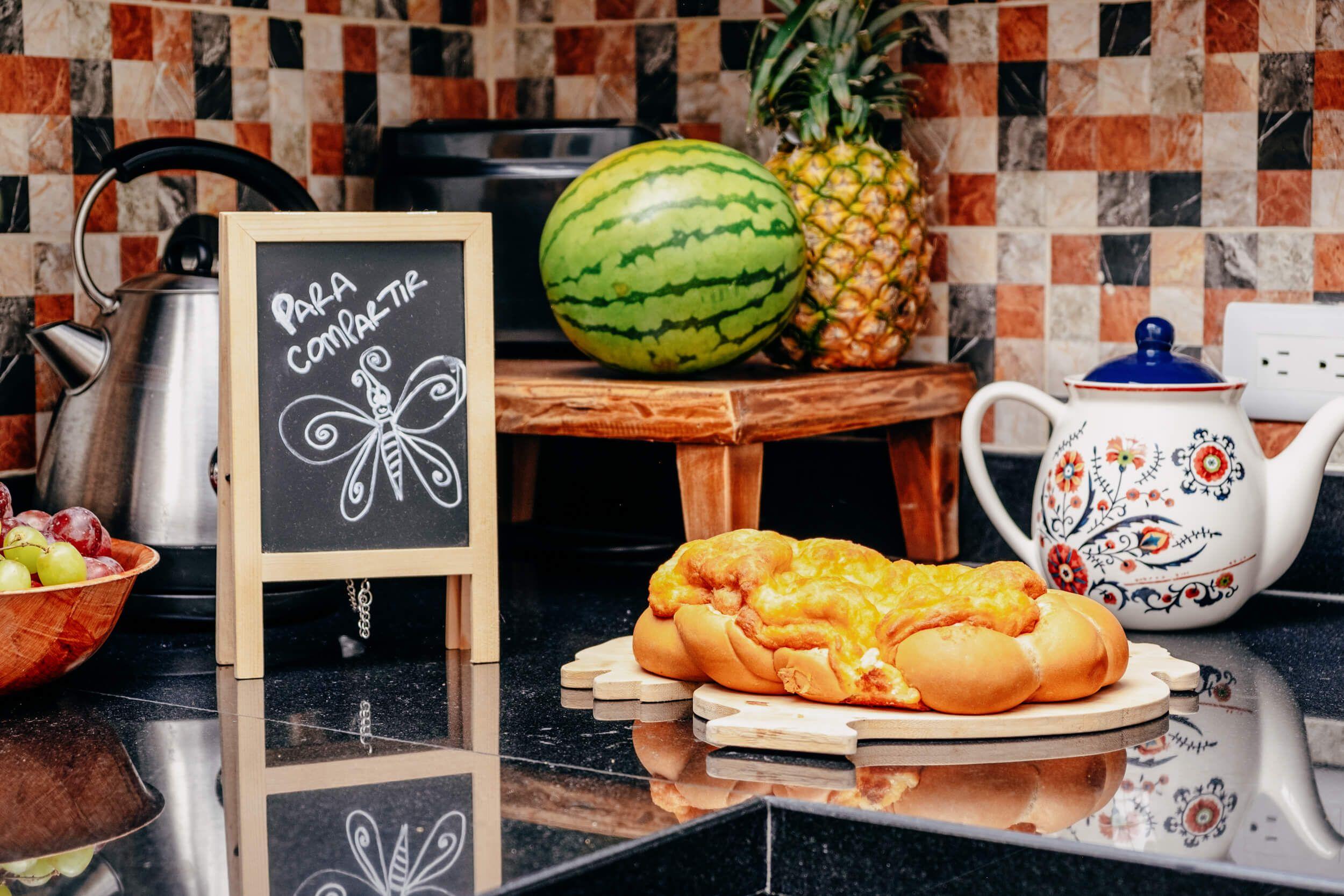 Shared kitchen counter with fresh fruit, bread, and a 'Para Compartir' (to share) chalkboard sign