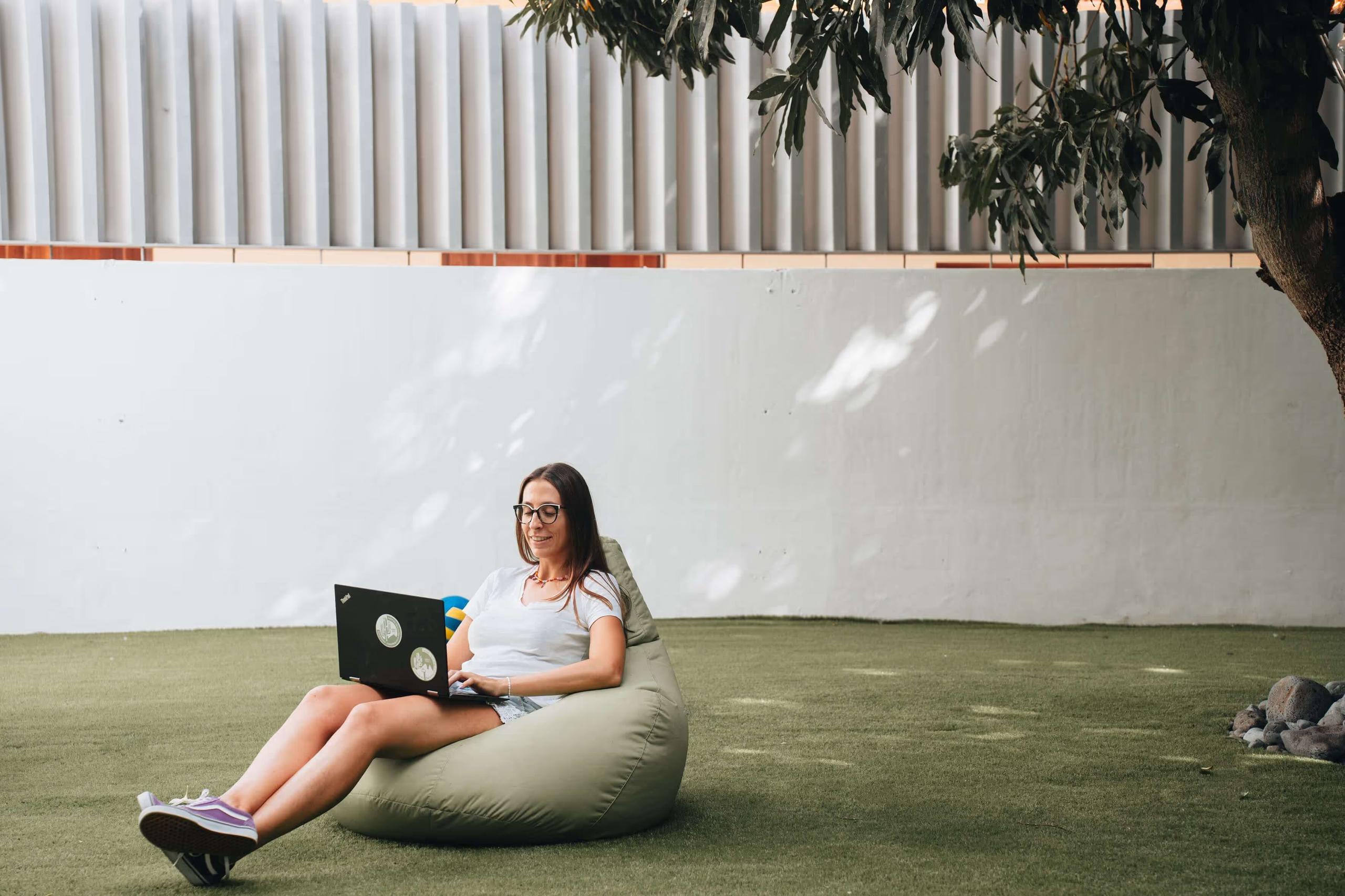 Person working on a laptop on a bean bag chair in the garden patio under a mango tree