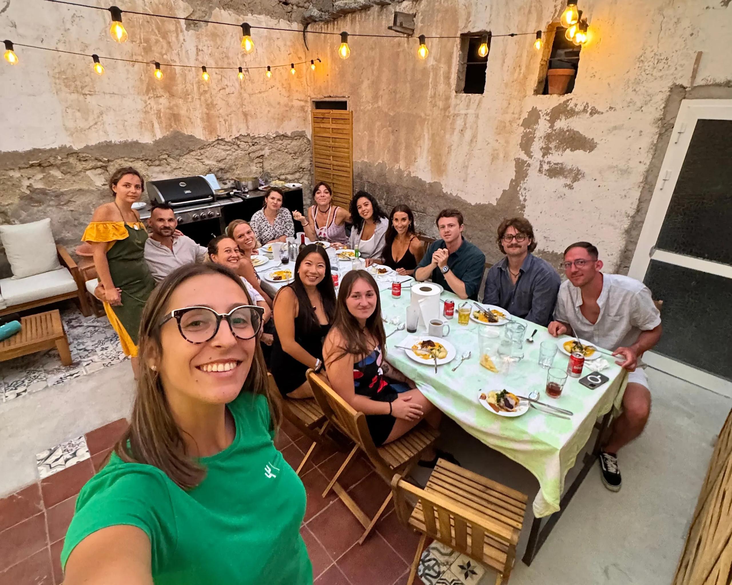 Large group of colivers sharing a dinner at a long table in the outdoor BBQ area, with string lights overhead and the BBQ grill visible in the background
