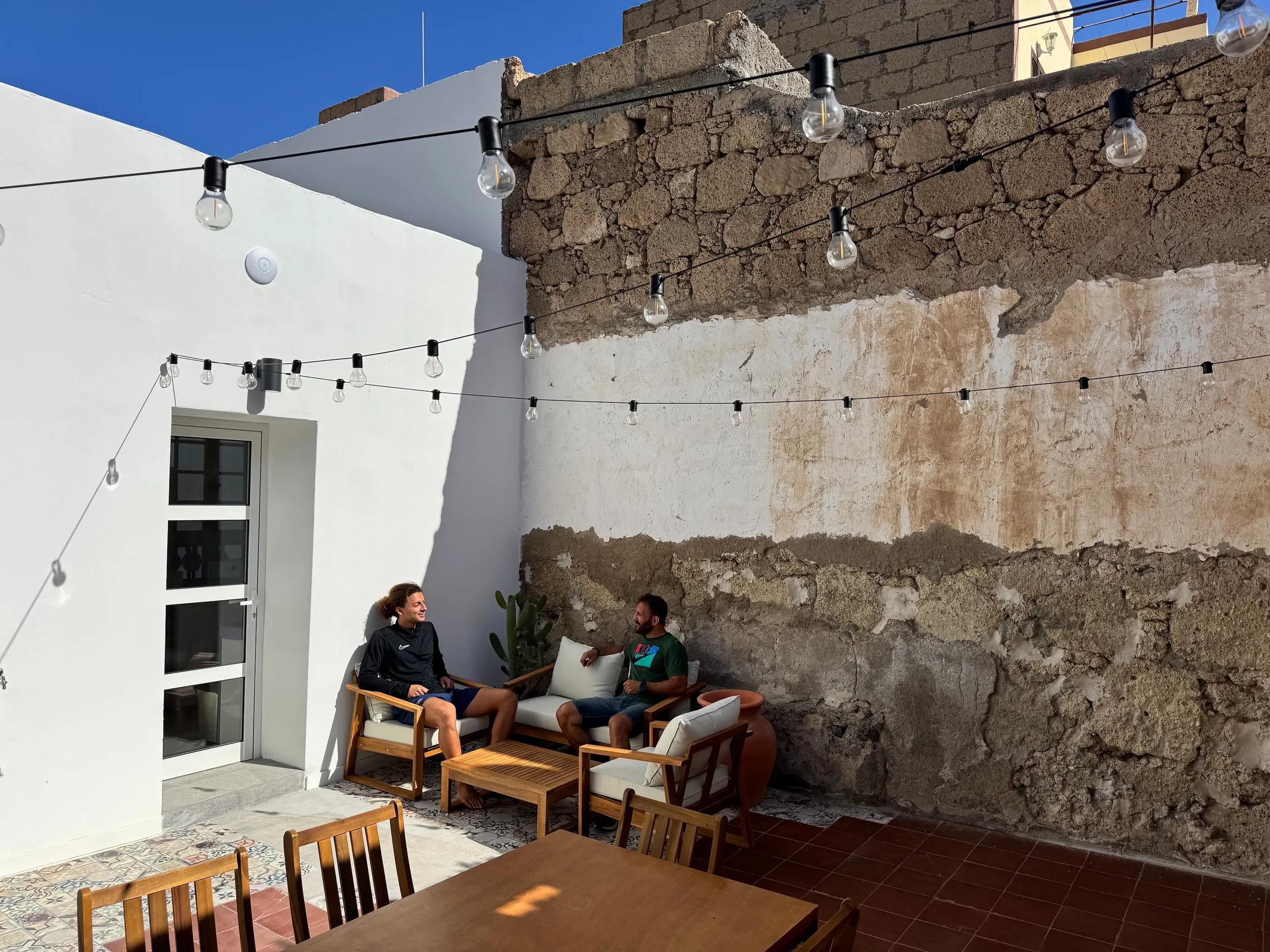 Outdoor patio with wooden armchairs, dining table and string lights, two colivers chatting