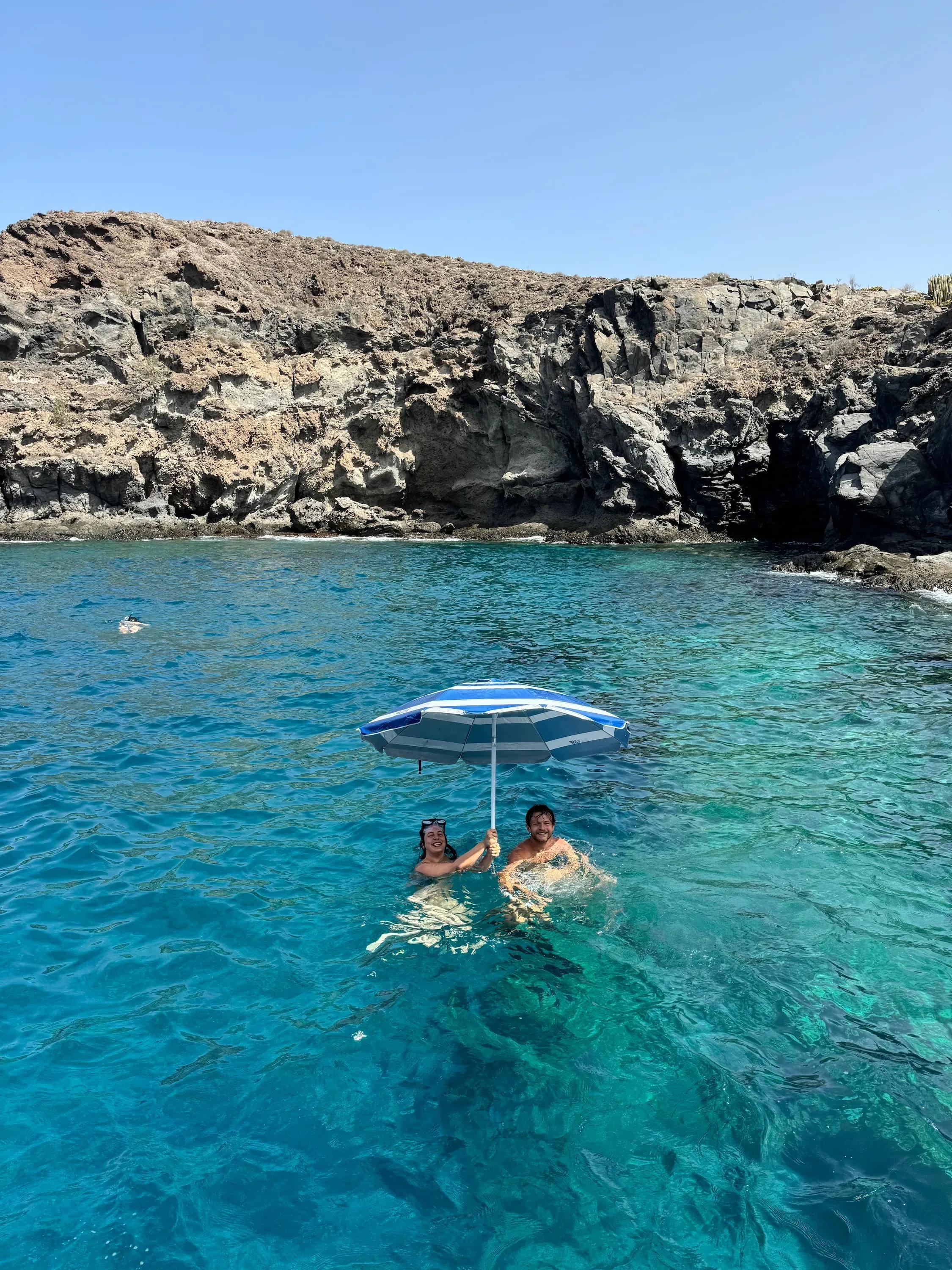 Two people swimming in clear turquoise water near rocky cliffs, holding a beach umbrella