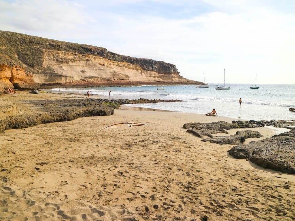 Sandy beach near Adeje with rocky cliffs, calm waves and sailboats in the background