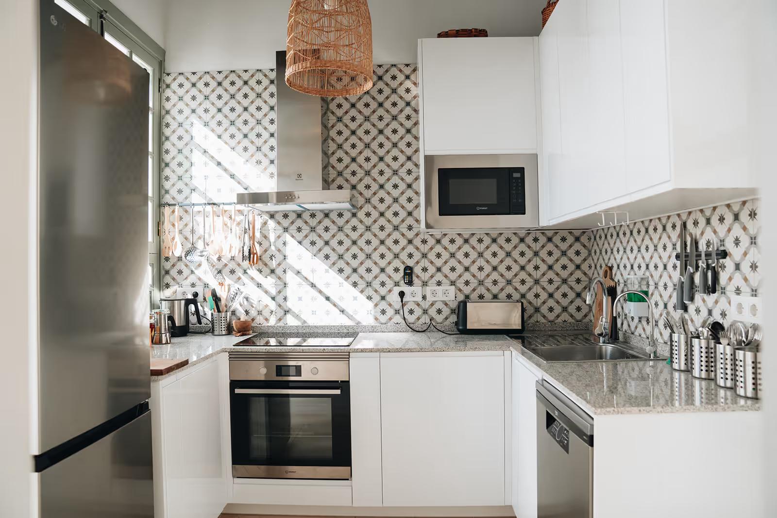 Fully equipped shared kitchen with white cabinets, patterned tile backsplash, stainless steel appliances, oven, microwave and wicker pendant light
