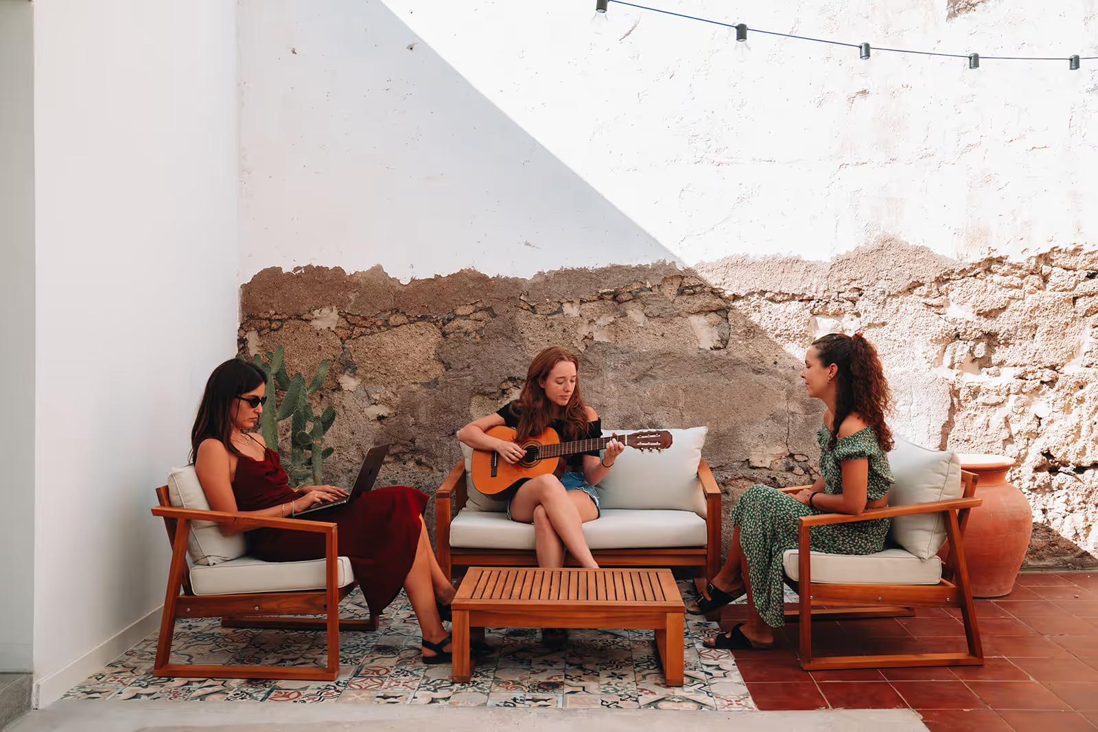 Three women relaxing on the outdoor patio — one playing guitar, one on a laptop, one chatting — surrounded by white walls and string lights