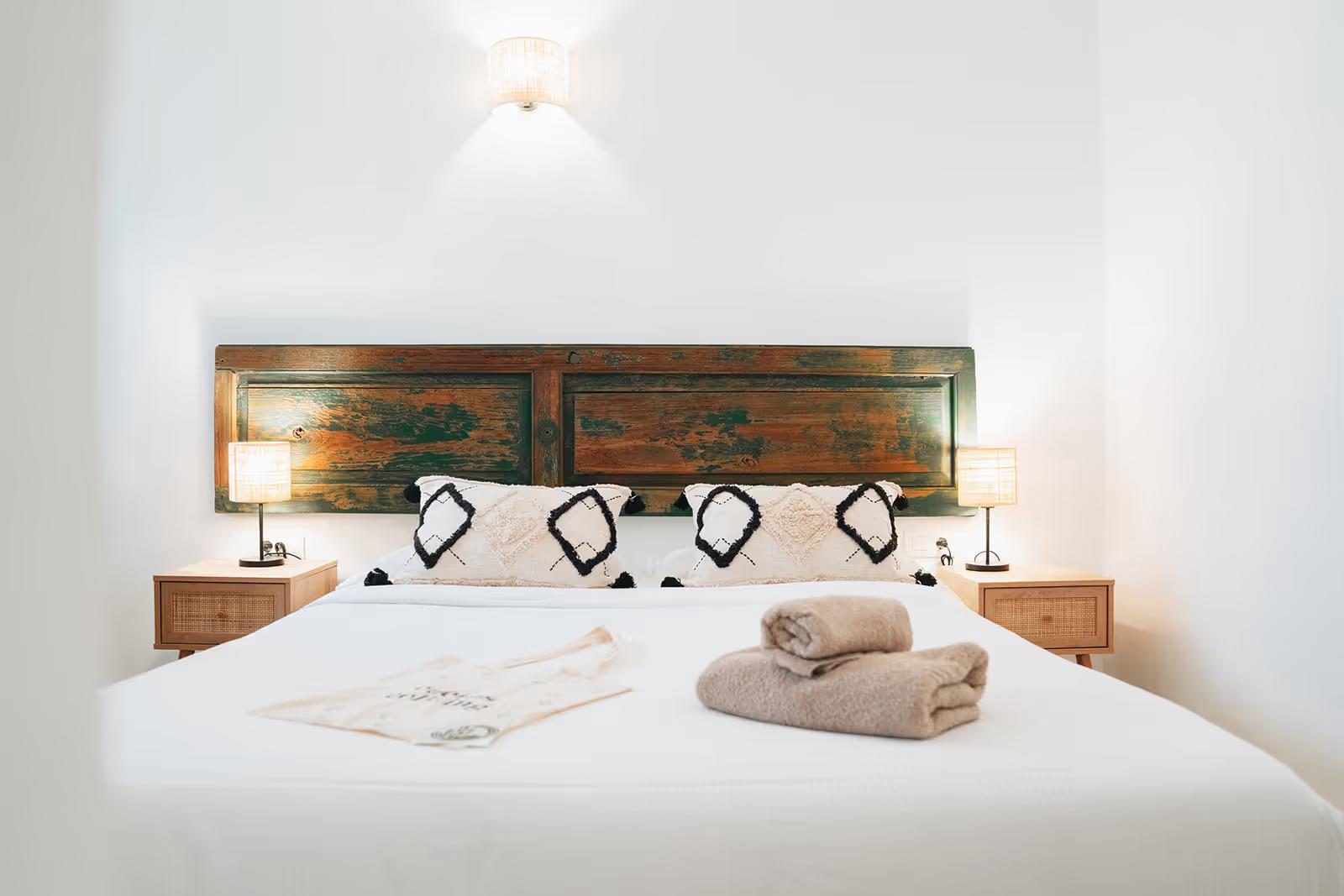 Bright double bedroom with a rustic painted wooden headboard, white bedding, folded towels and two bedside lamps