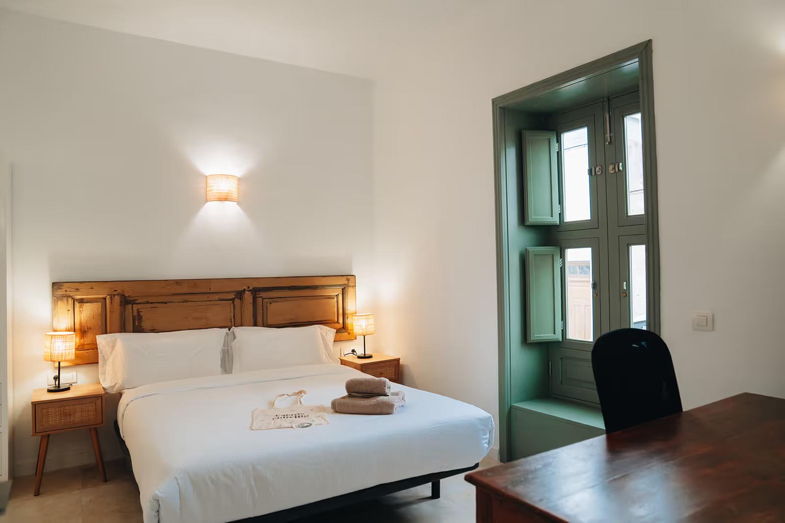 Spacious double bedroom with wooden bed, white bedding, traditional green-framed window and wooden desk