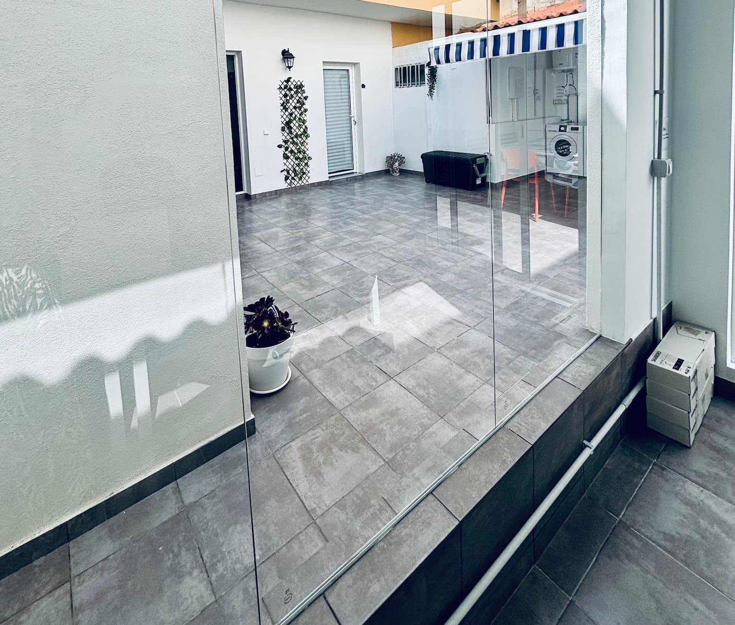Spacious tiled outdoor terrace/patio area viewed through glass panels, with plants and retractable awning visible