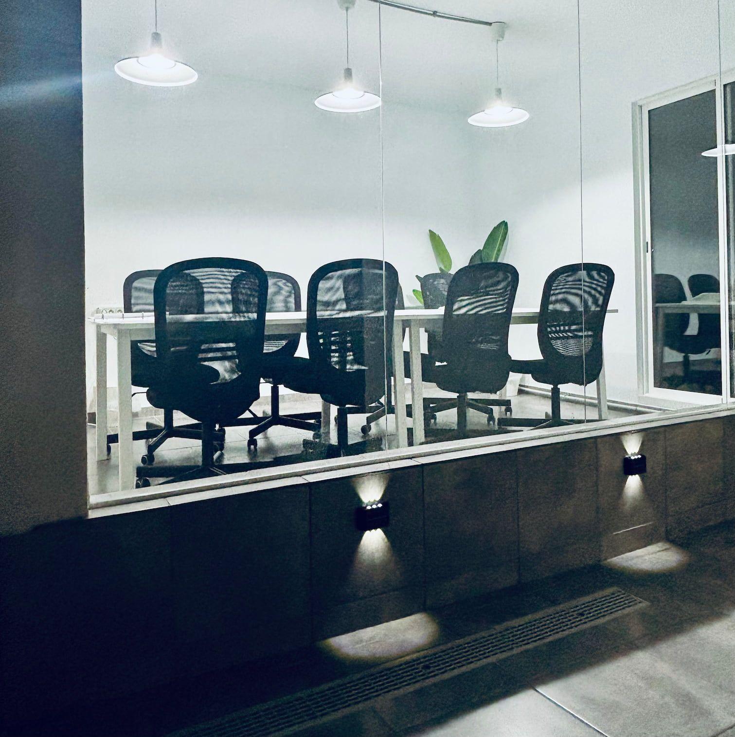 Glass-enclosed coworking room with long white desks and black mesh chairs, pendant lighting