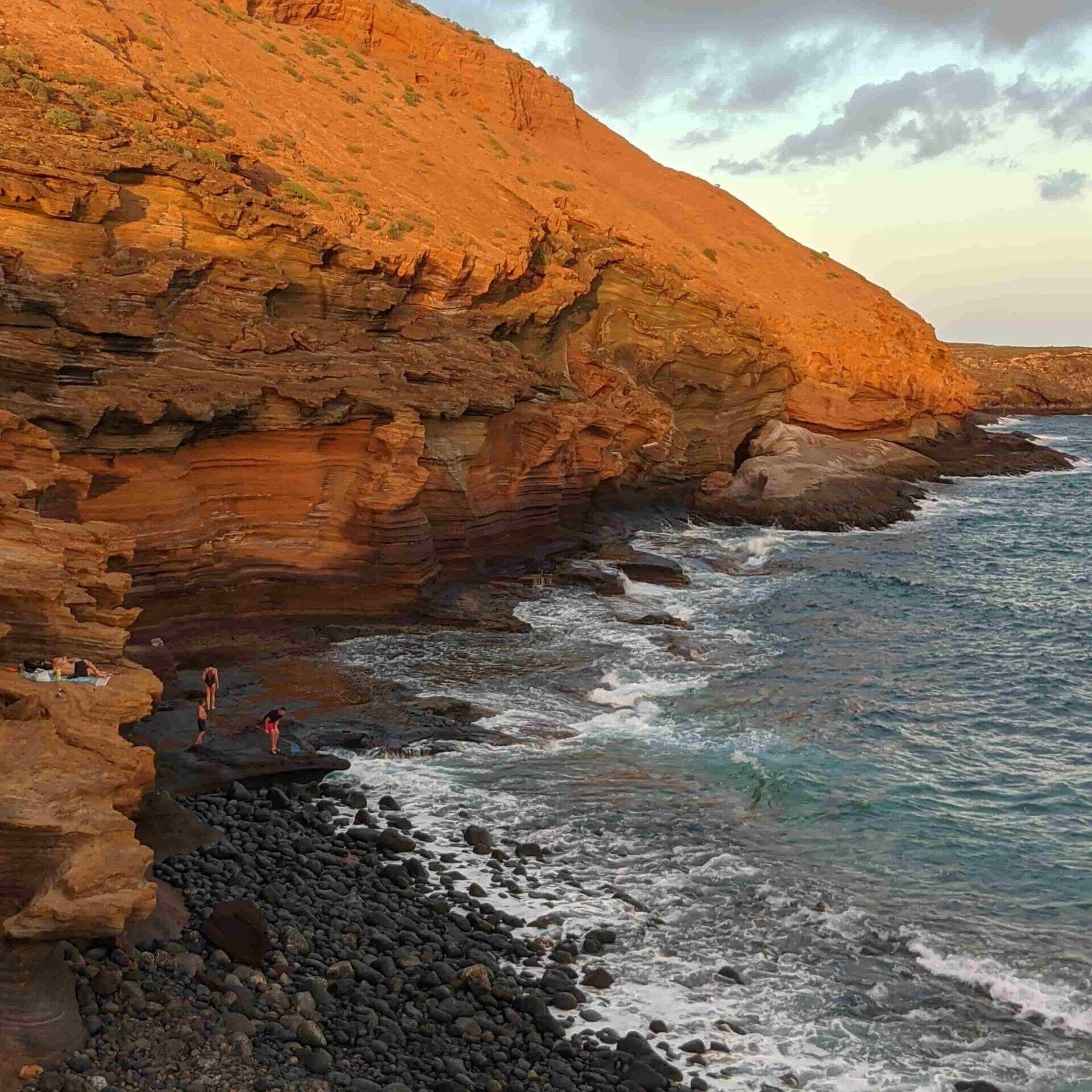 Volcanic red rock cliffs meeting the Atlantic Ocean at a black pebble beach in Tenerife, with people exploring at sunset