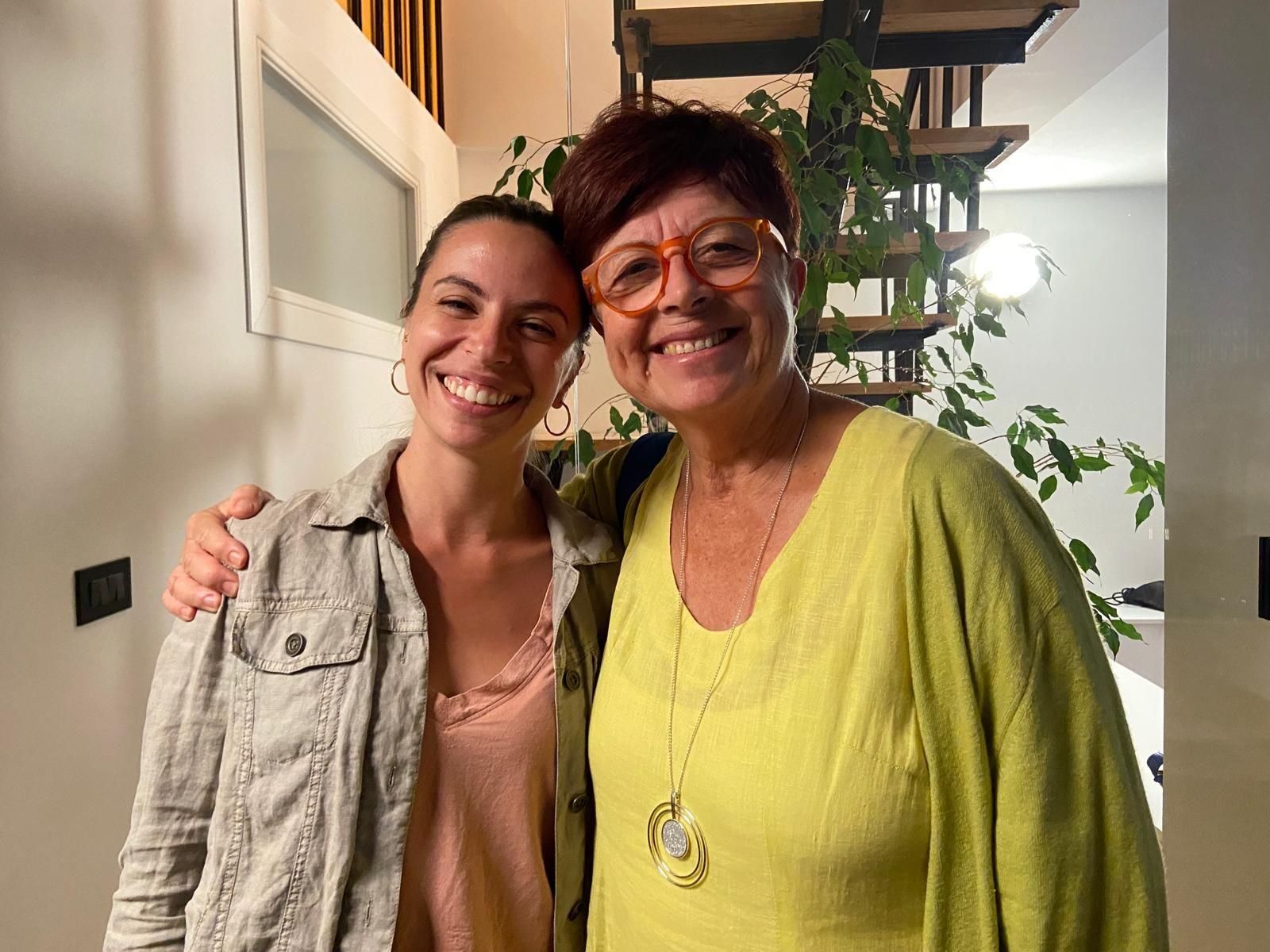 Two women smiling together inside Casa Netural coliving space, with a staircase and plants in the background