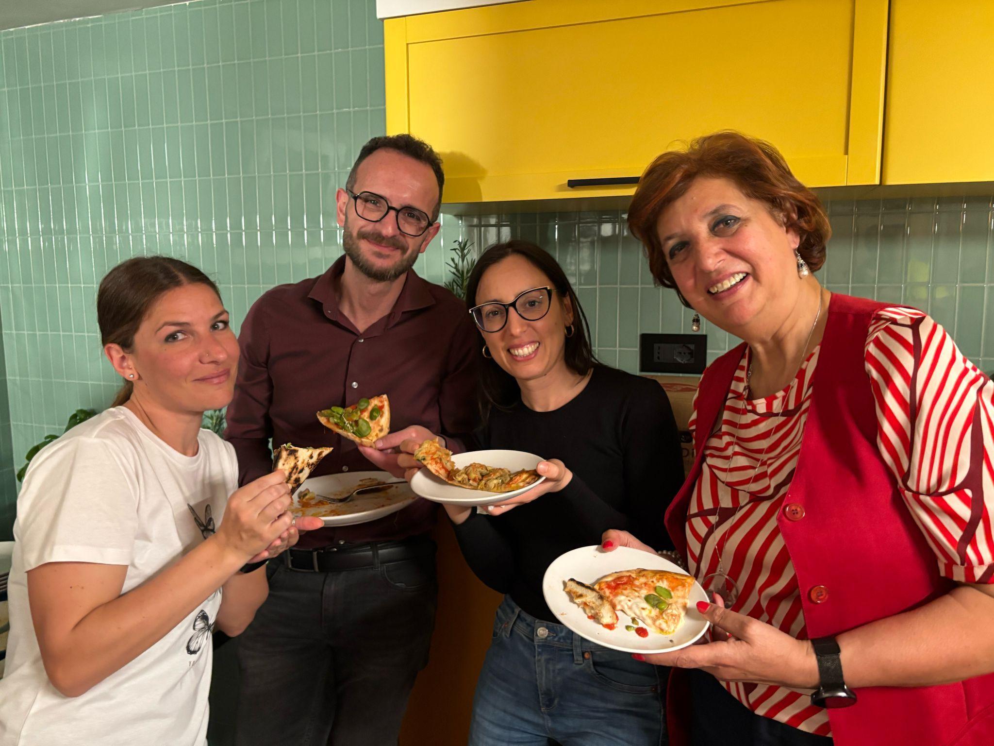 Four people smiling and holding plates of food in the kitchen during a community dinner at Casa Netural Matera