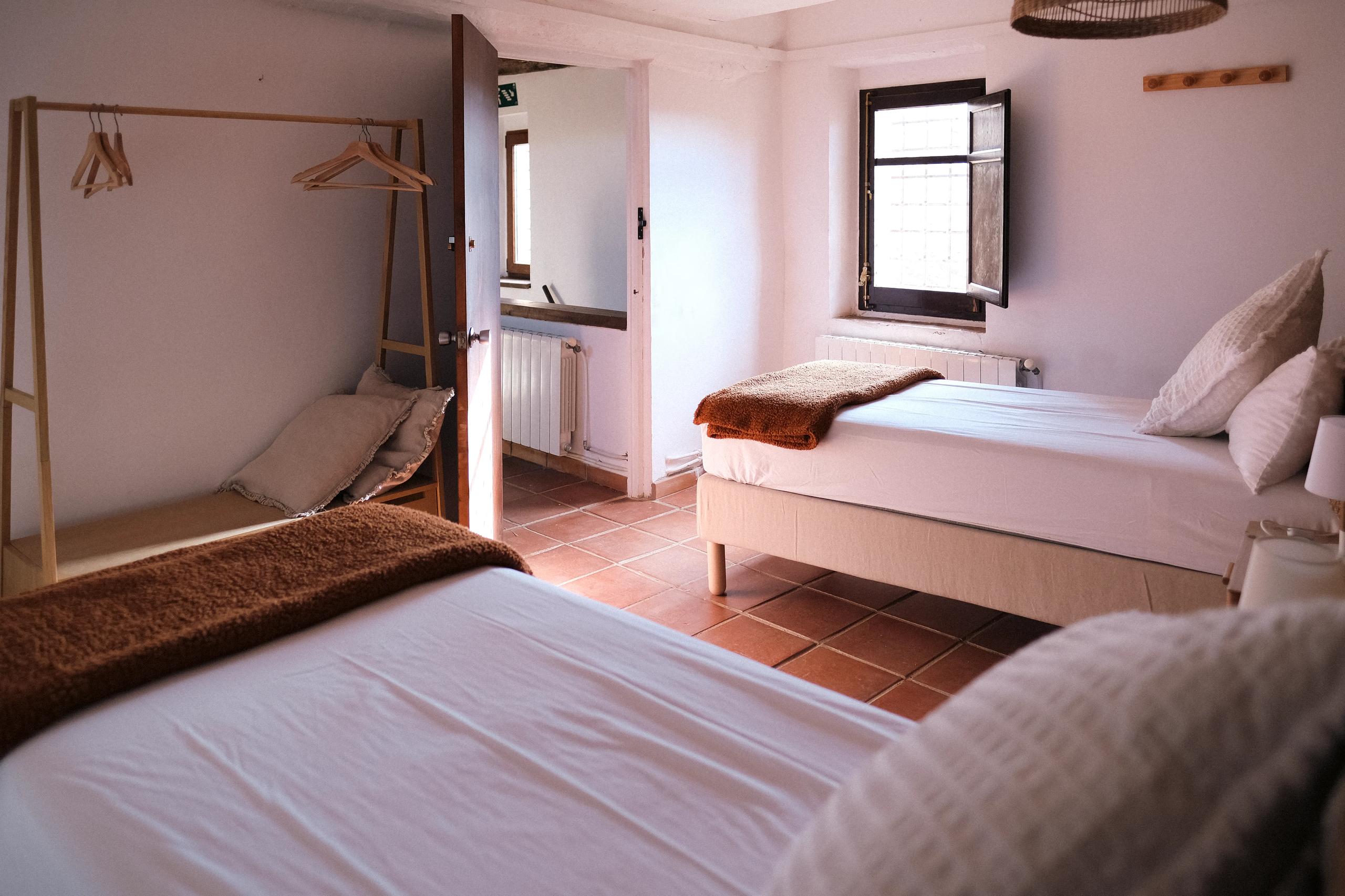 A shared twin room with two single beds, wooden clothes rack, and terracotta tile floor