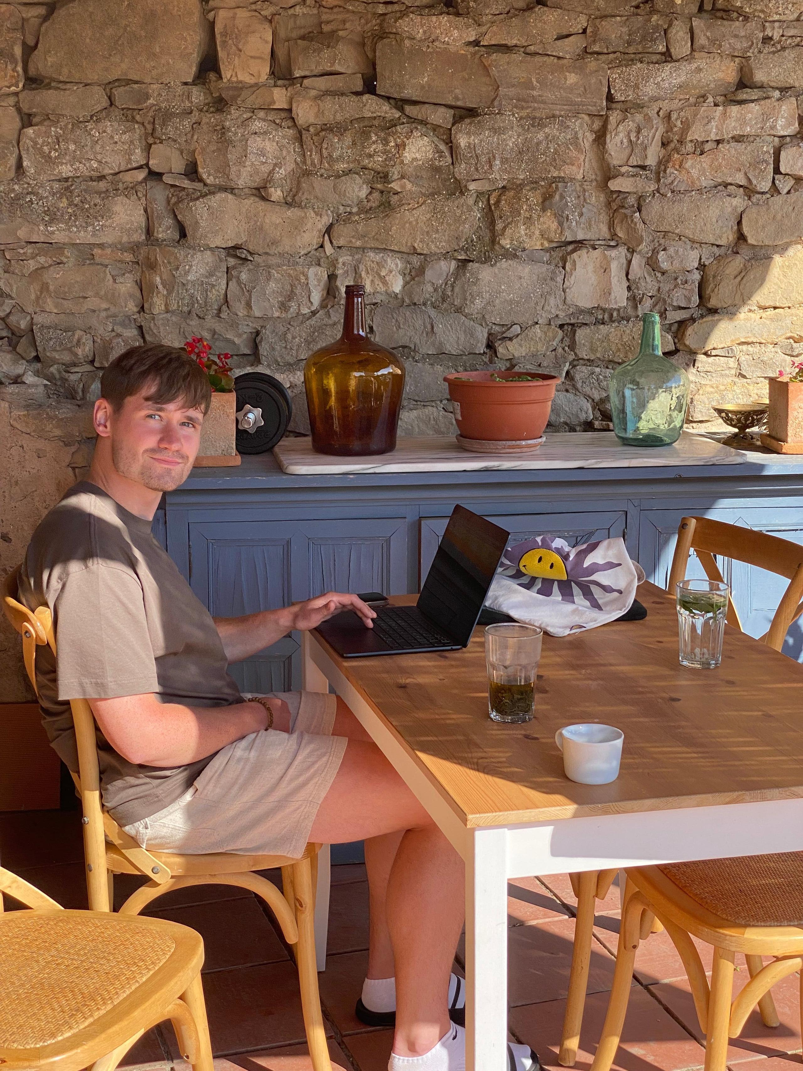 A person working on a laptop at an outdoor table next to a stone wall