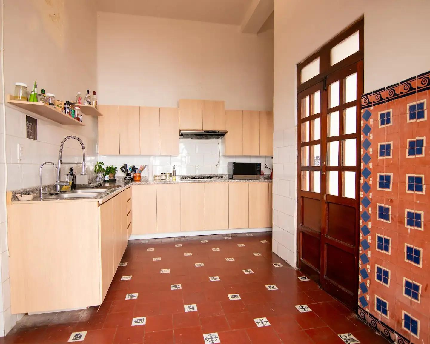 Fully equipped shared kitchen with light wood cabinets, countertops, sink, microwave, and traditional tiled floor