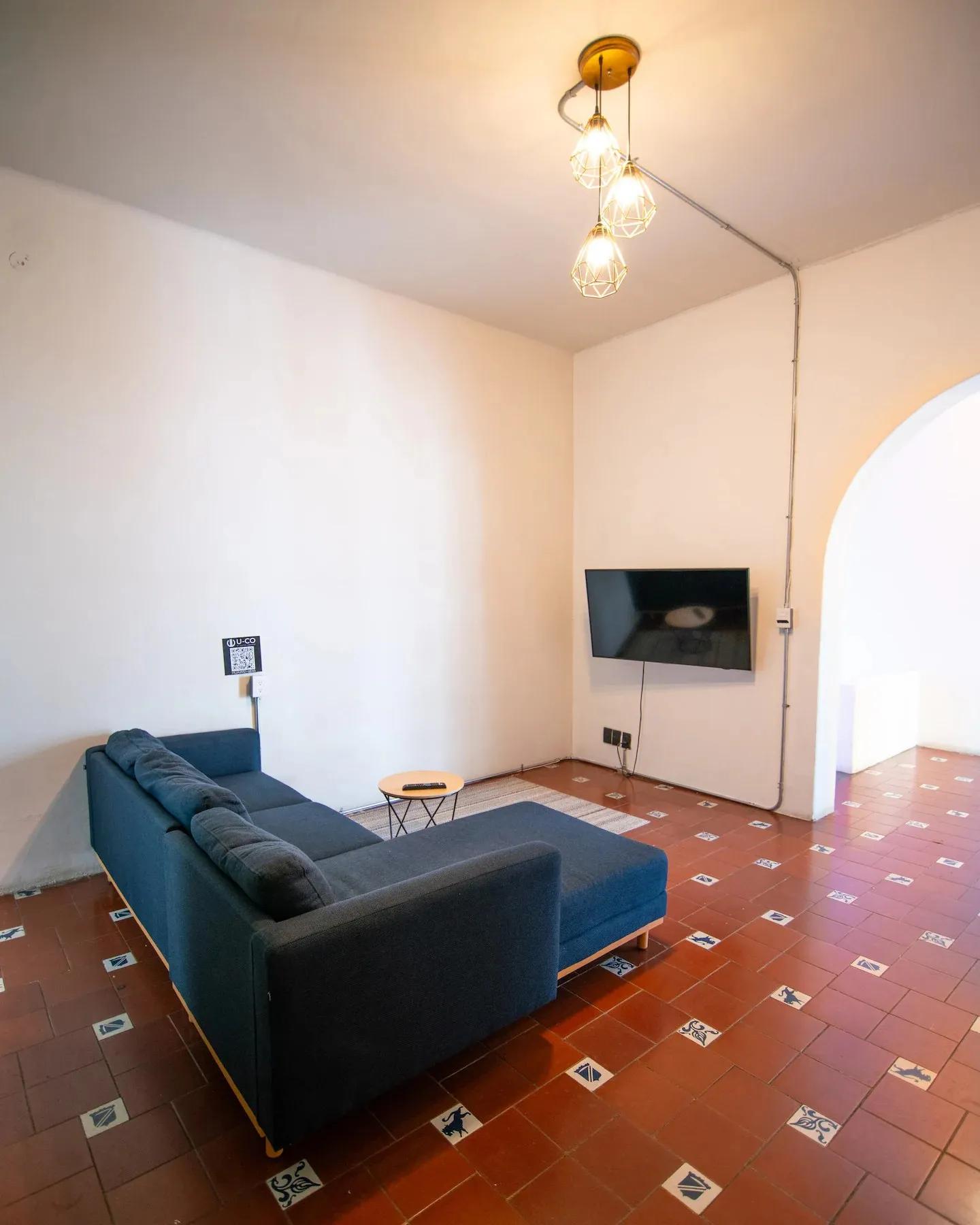Living room with a large blue sectional sofa, wall-mounted TV and decorative pendant lights on terracotta tiled floor