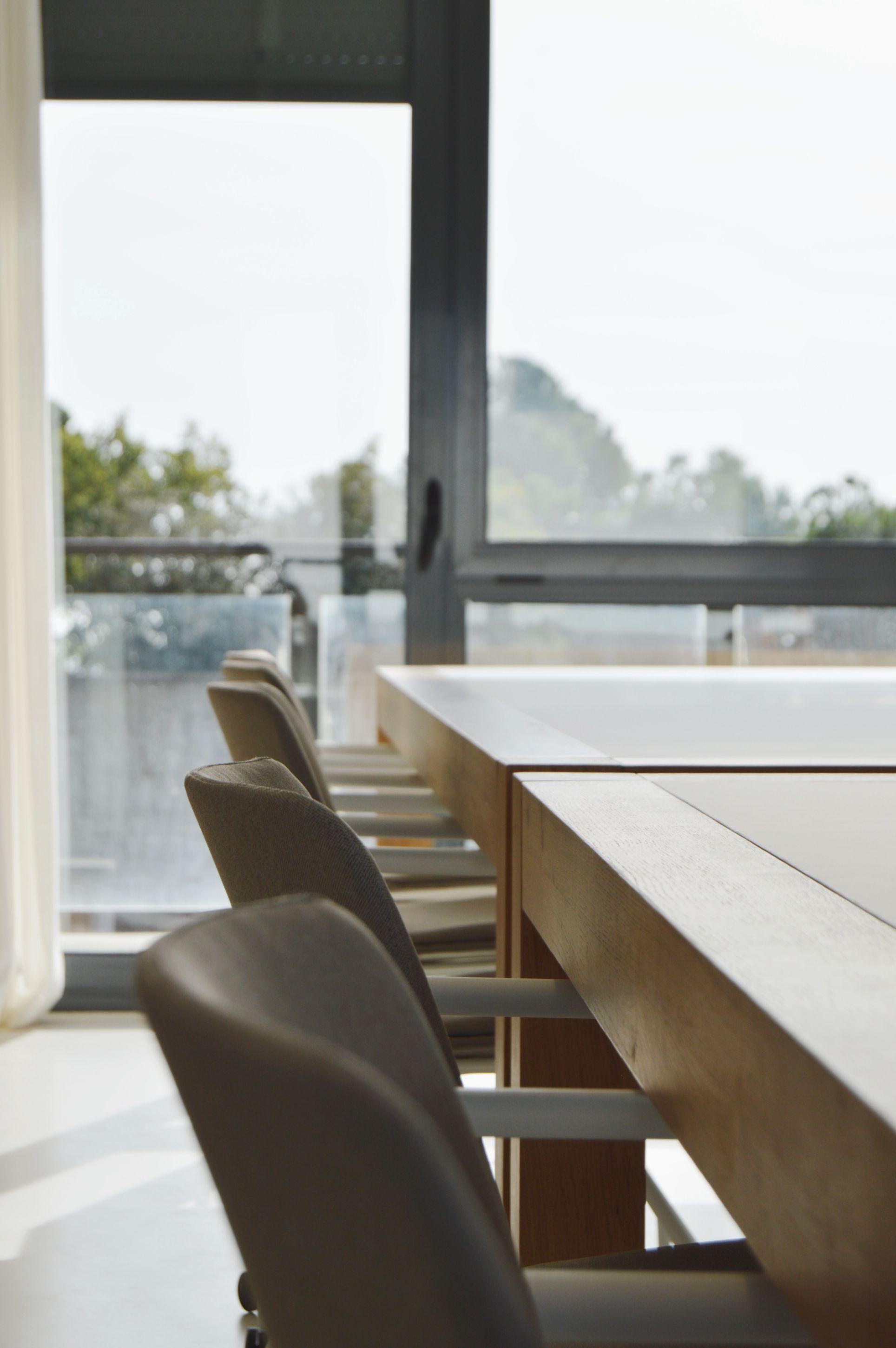 Close-up of a row of coworking desks with chairs, viewed from the side, with large windows and mountain views beyond.