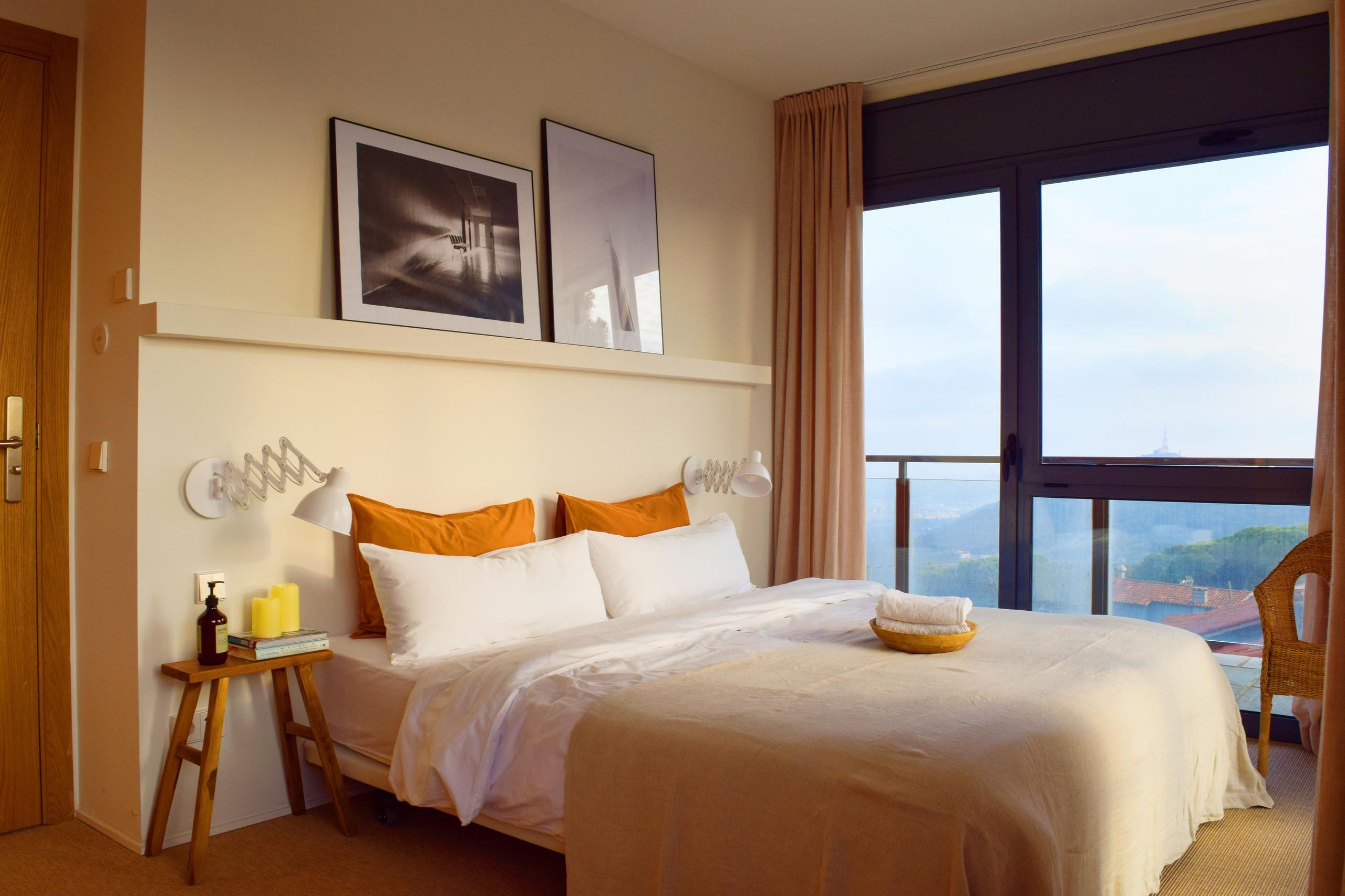 King-size bed with white and orange bedding, framed artwork above the headboard, and a large window with mountain views and a private terrace.