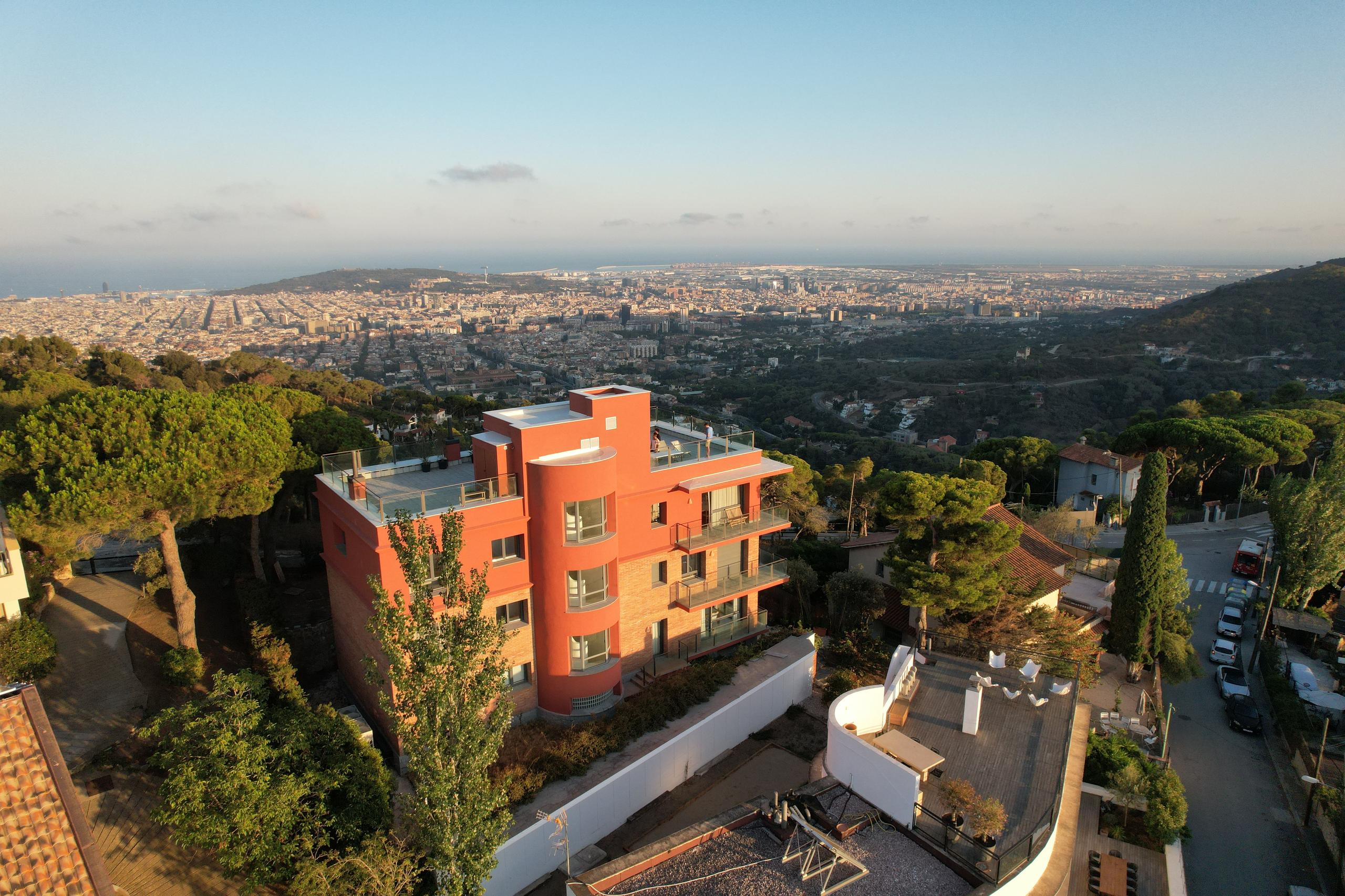 Aerial drone shot of the Circles House Barcelona building, a red/terracotta multi-storey structure set in a natural park above Barcelona, with panoramic city and sea views in the background.