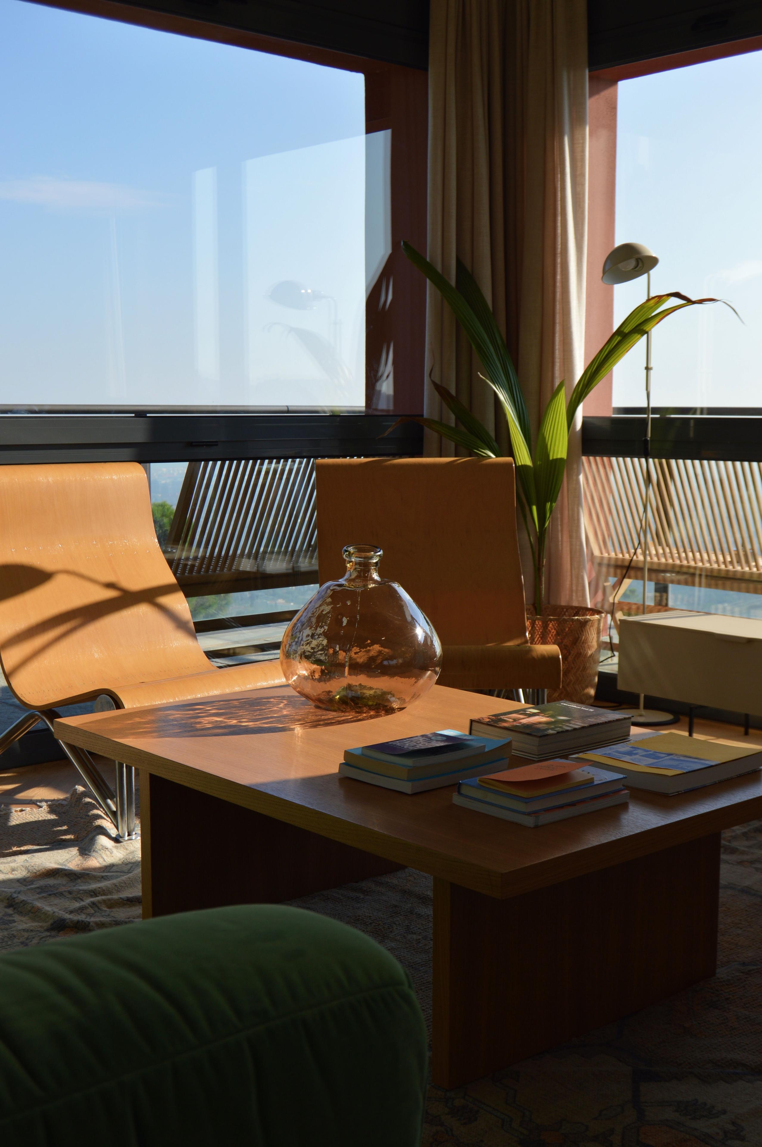 Warm, sunlit living room with a wooden coffee table, books, a glass vase, plants, and large panoramic windows overlooking the sea.