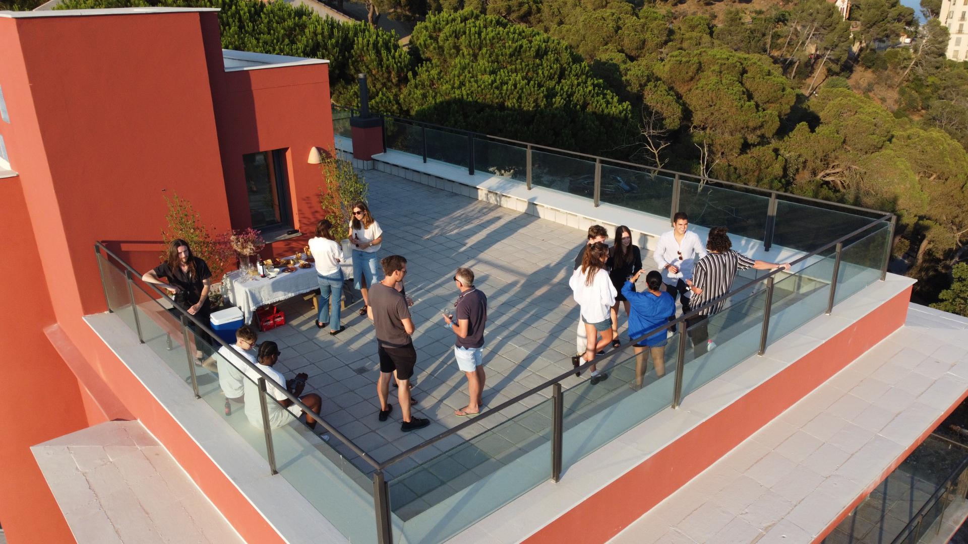 Aerial view of a group of people gathered on the rooftop terrace for a social event, with the red building facade and surrounding trees visible.