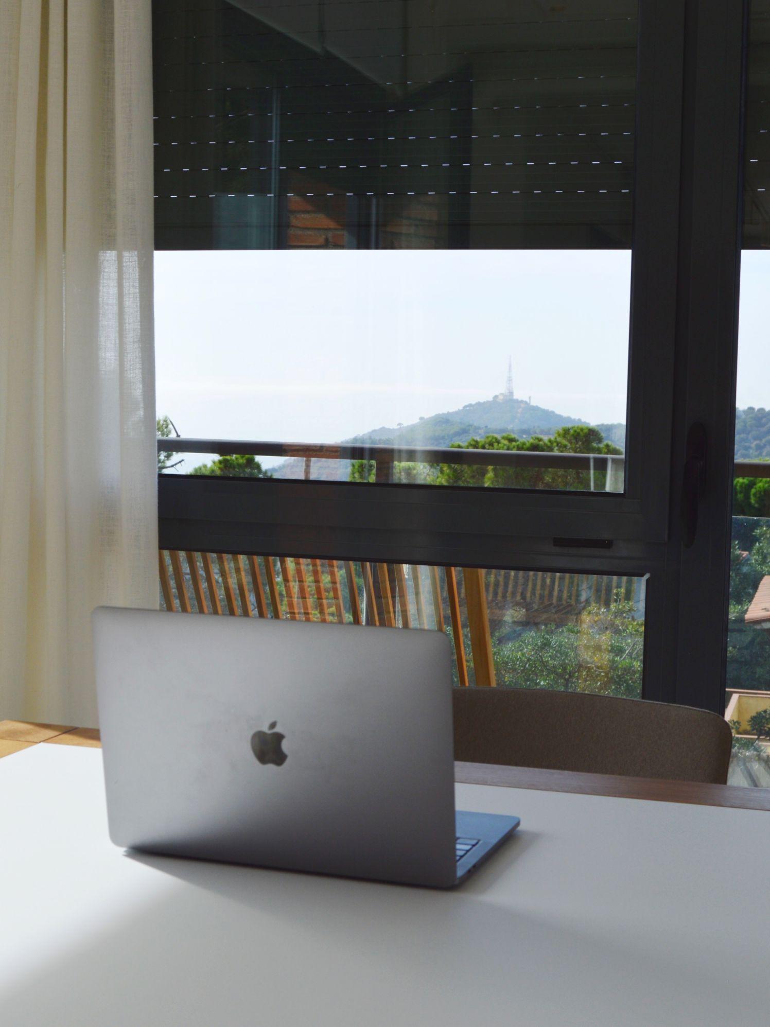 A MacBook laptop open on a white coworking desk, with a mountain view through the window behind.