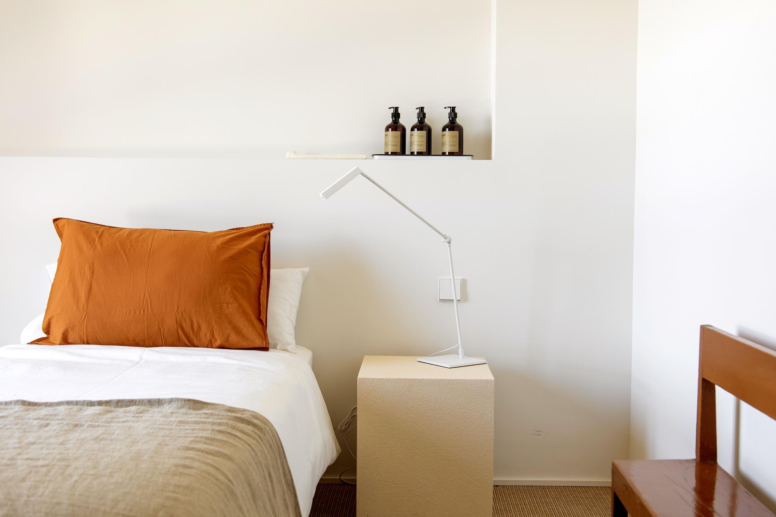 King studio room with a bed featuring an orange pillow, a bedside lamp, and toiletries on a shelf above.
