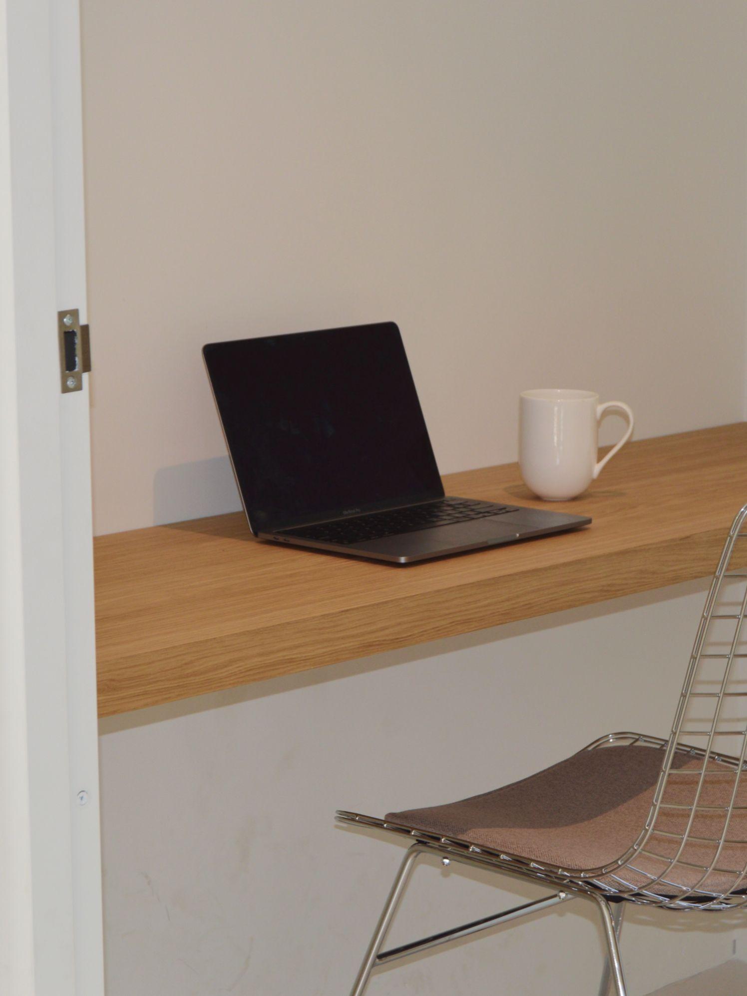 A laptop and coffee mug on a wooden floating desk inside a private cabin, with a wire chair.