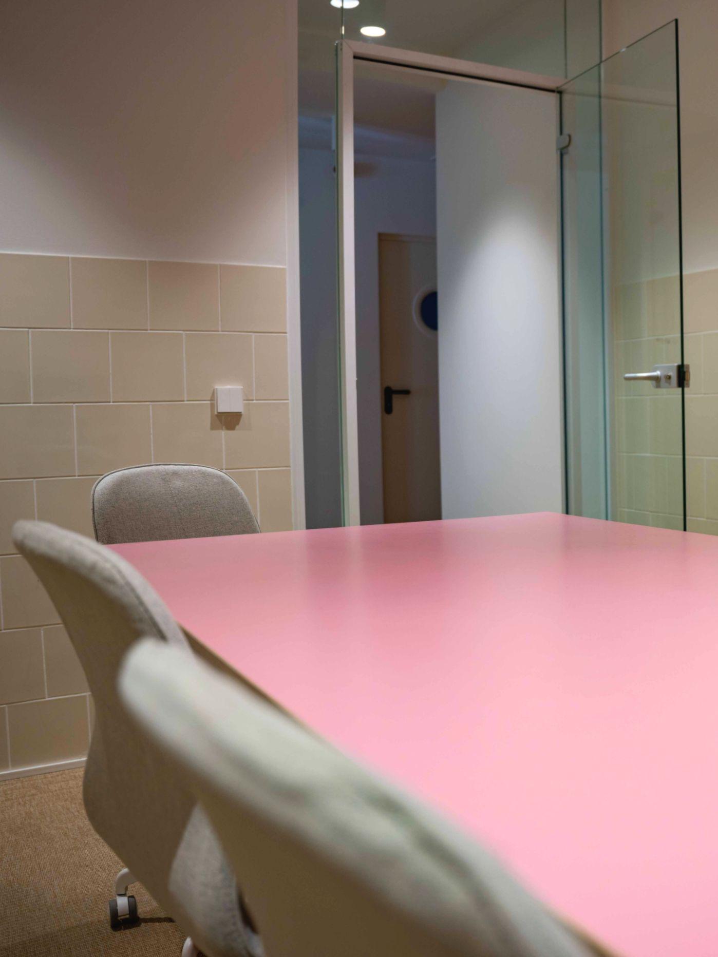 View of the meeting room through its glass door, showing the pink table and white chairs inside.