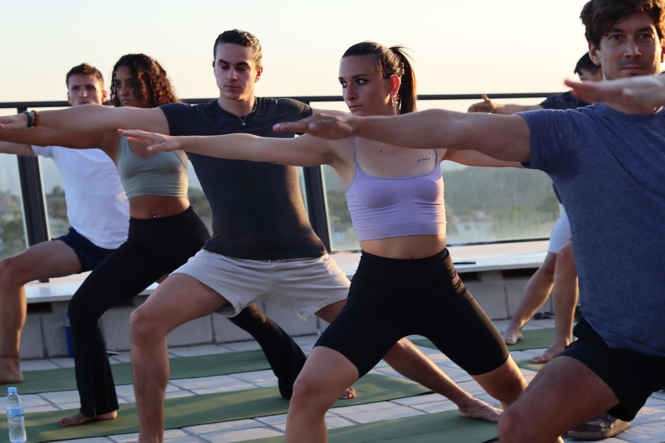 Group yoga class on the rooftop terrace at sunset, with participants in warrior pose.