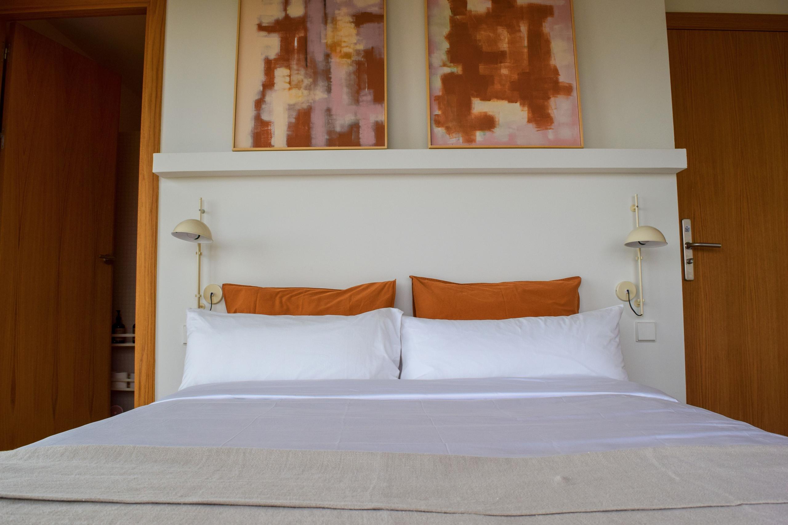 Close-up of a king-size bed with white and orange pillows, wall sconces, and abstract artwork above the headboard.