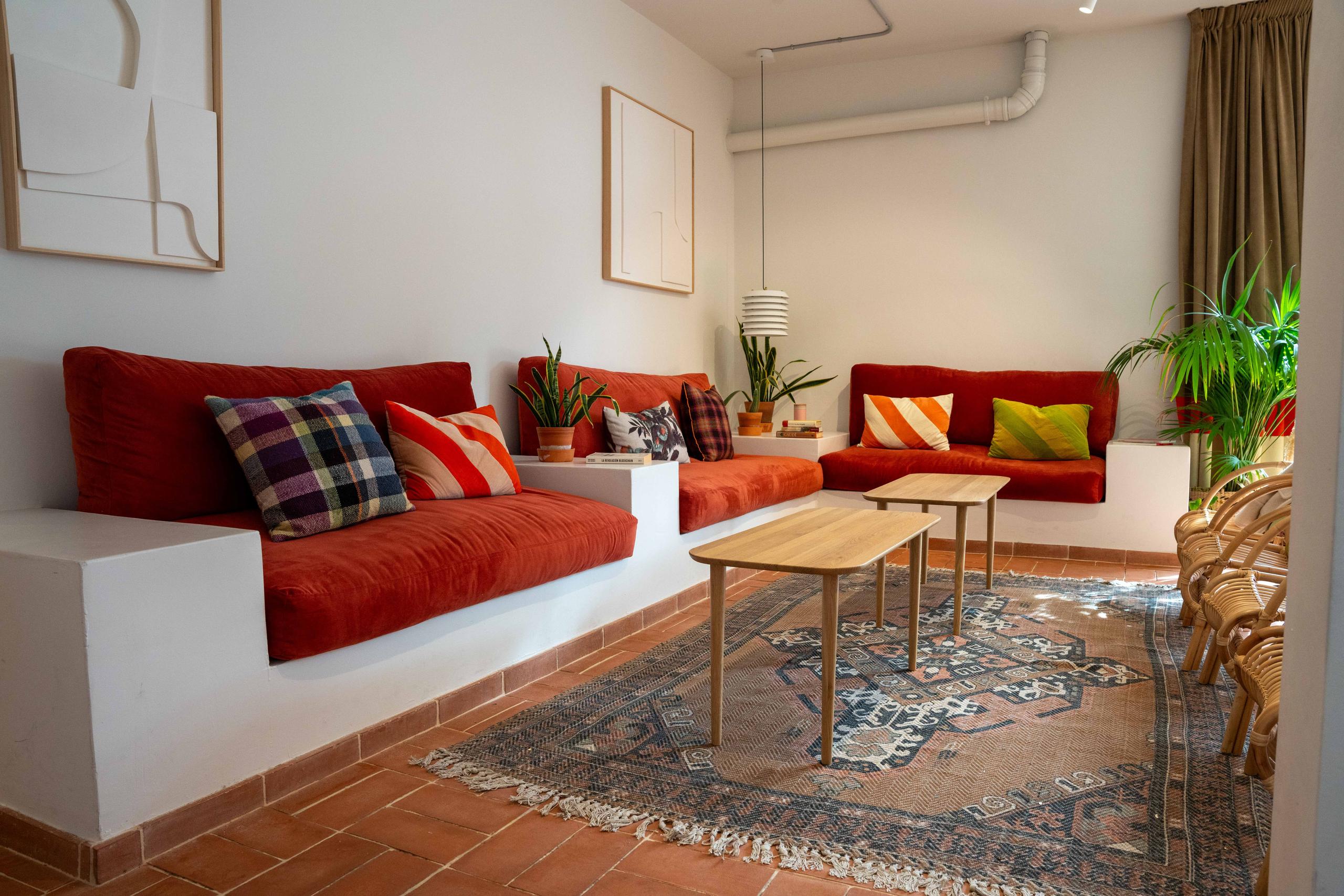 Cozy social club space with terracotta-red sofas, colourful cushions, a wooden coffee table, and plants on a terracotta tile floor.