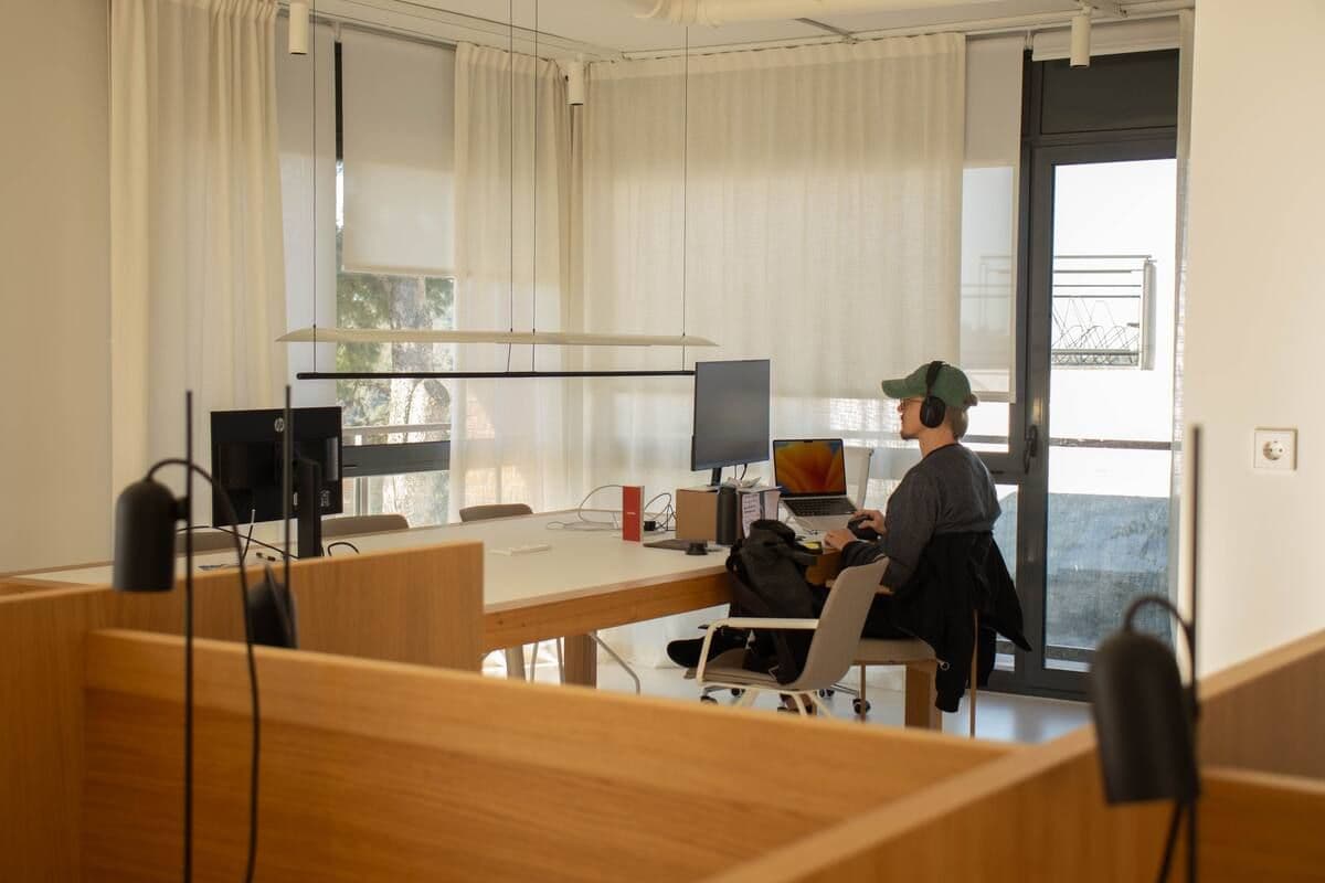 A person working at a desk in the bright coworking space, with monitors, sheer curtains, and a terrace door visible.